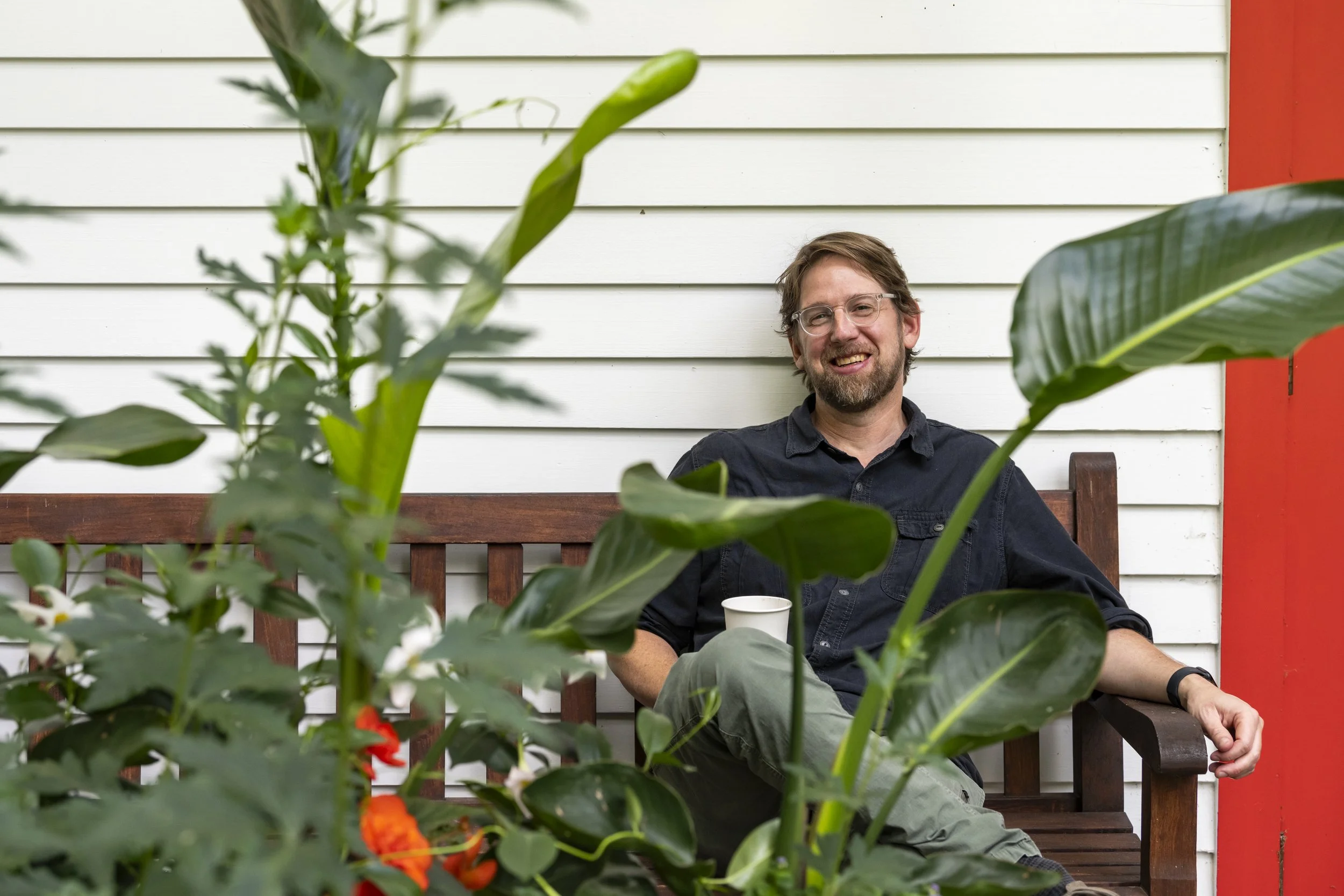 A man with glasses and a beard, smiling, seated on a wooden bench behind green plants, beside a white house wall with red trim, holding a white cup