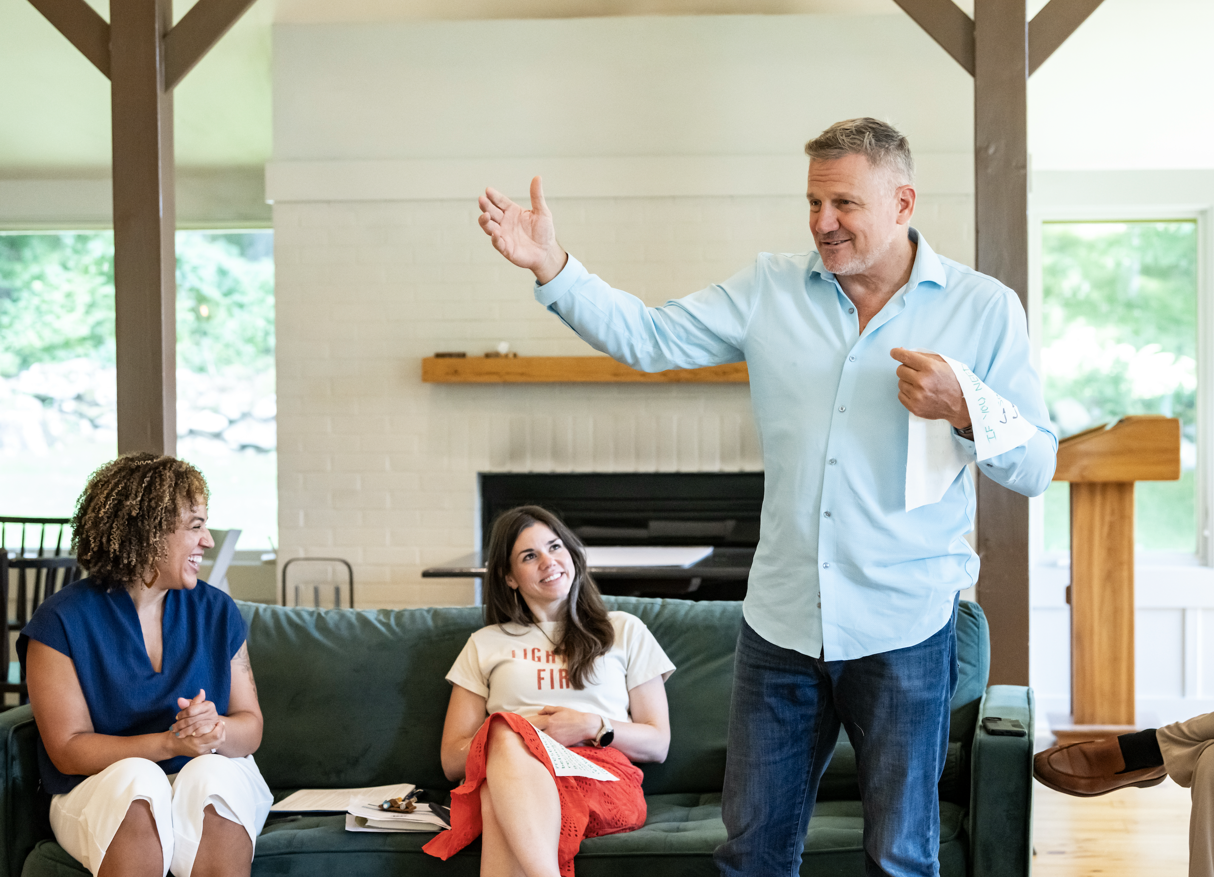 A group of people sitting on a green couch in a bright room, engaged in conversation. A man with short gray hair, wearing a light blue shirt, is standing and speaking to the group. Two women, one with curly hair and the other with straight hair, are smiling and listening.