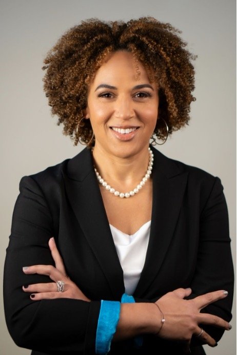 A woman with curly brown hair wearing a black blazer with turquoise cuffs, a white top, a pearl necklace, and rings, standing with arms crossed against a gray background.