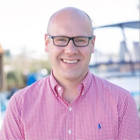 A smiling man wearing glasses and a red checkered button-up shirt, standing outdoors with boats in the background.
