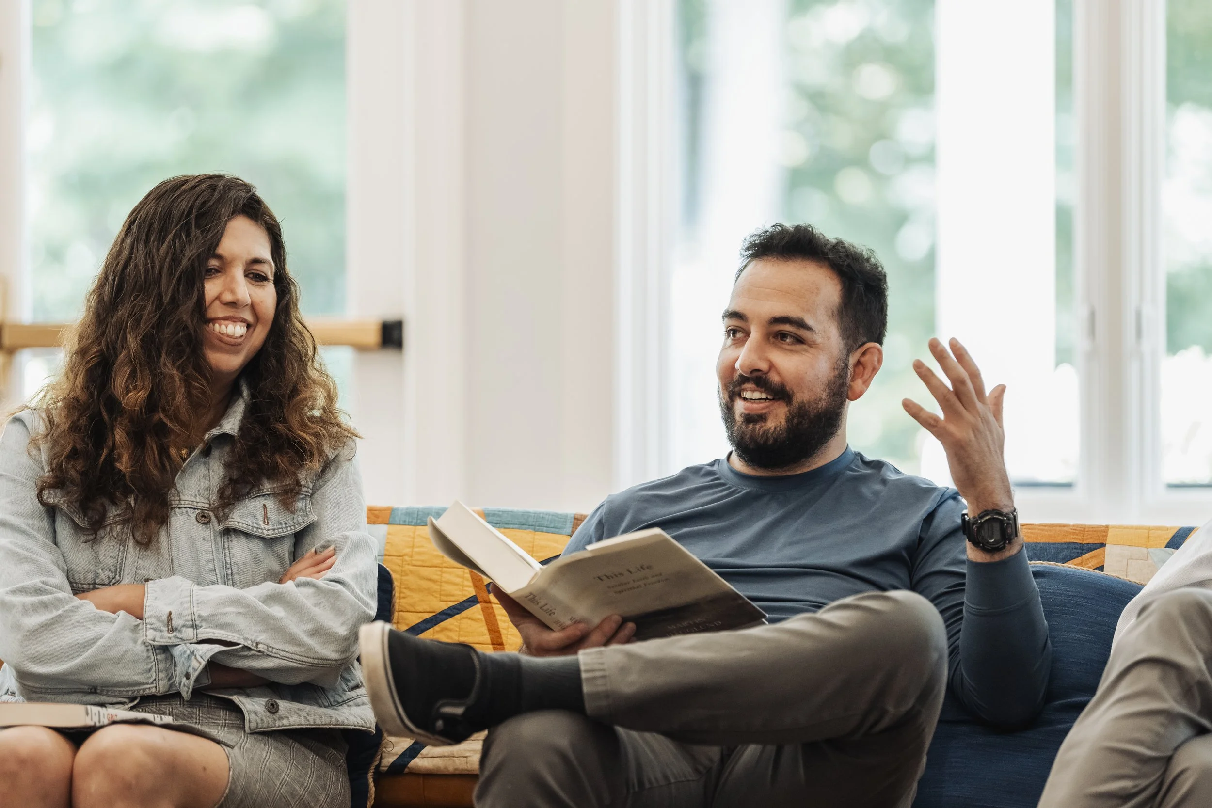 Two people sitting on a couch in a bright room, engaged in conversation. The woman on the left has long, curly brown hair and is smiling, wearing a light denim jacket. The man on the right has short, dark hair, a beard, and is gesturing with his right hand while holding an open book in his left hand, wearing a dark blue t-shirt.