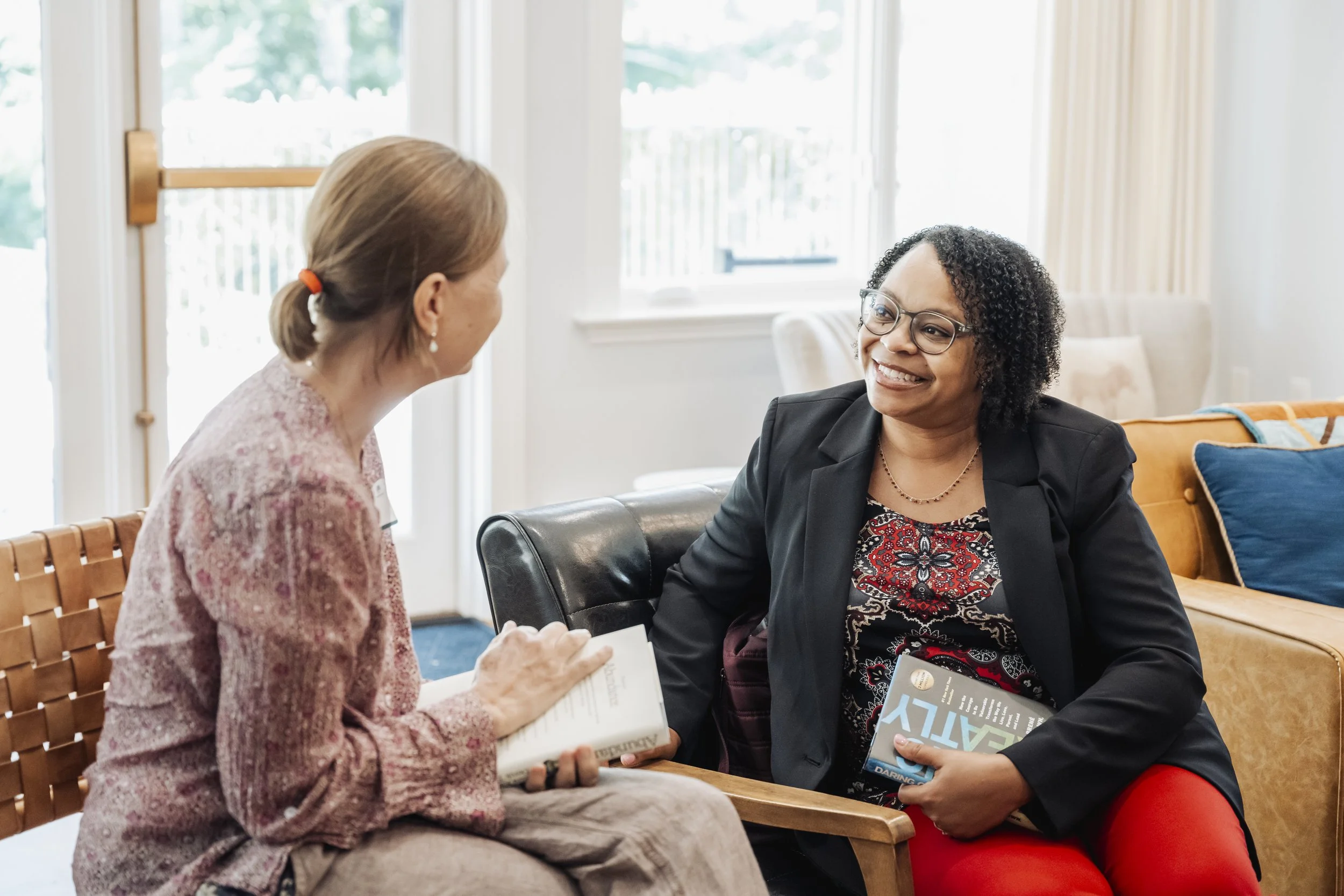 Two women sitting and talking inside a bright room with large windows. One woman has red hair tied back and is wearing a pink patterned blouse, holding a book. The other woman has dark curly hair, glasses, wearing a dark blazer, red pants, and holding a magazine. They are smiling and engaging in a friendly conversation.
