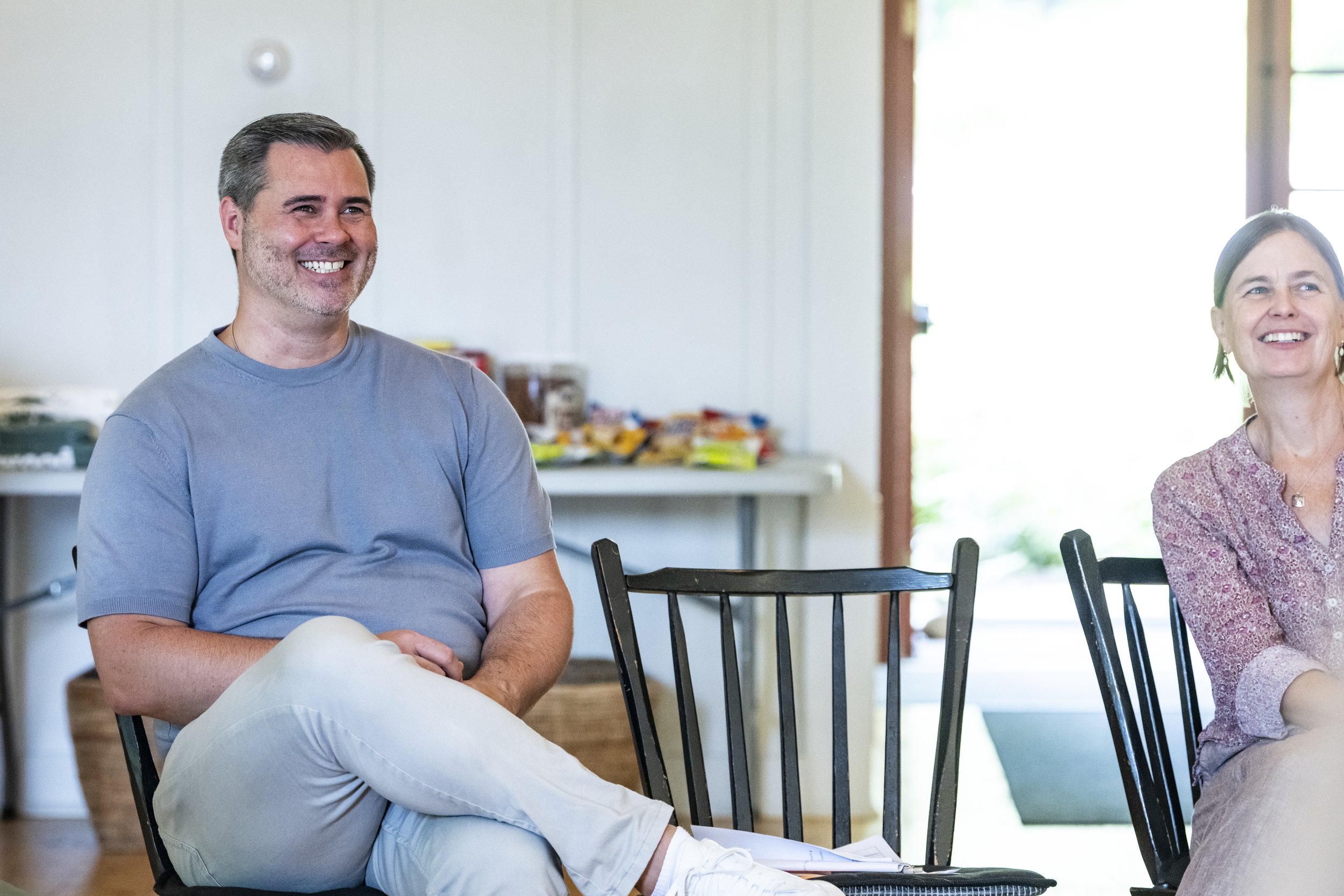 A man and a woman sitting on chairs in a brightly lit room, smiling and engaged in conversation.