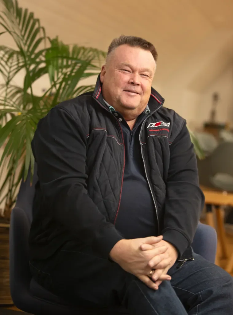 Mies istuu tuolilla ja hymyilee kuvassa, taustalla kasvi ja huonekalut.