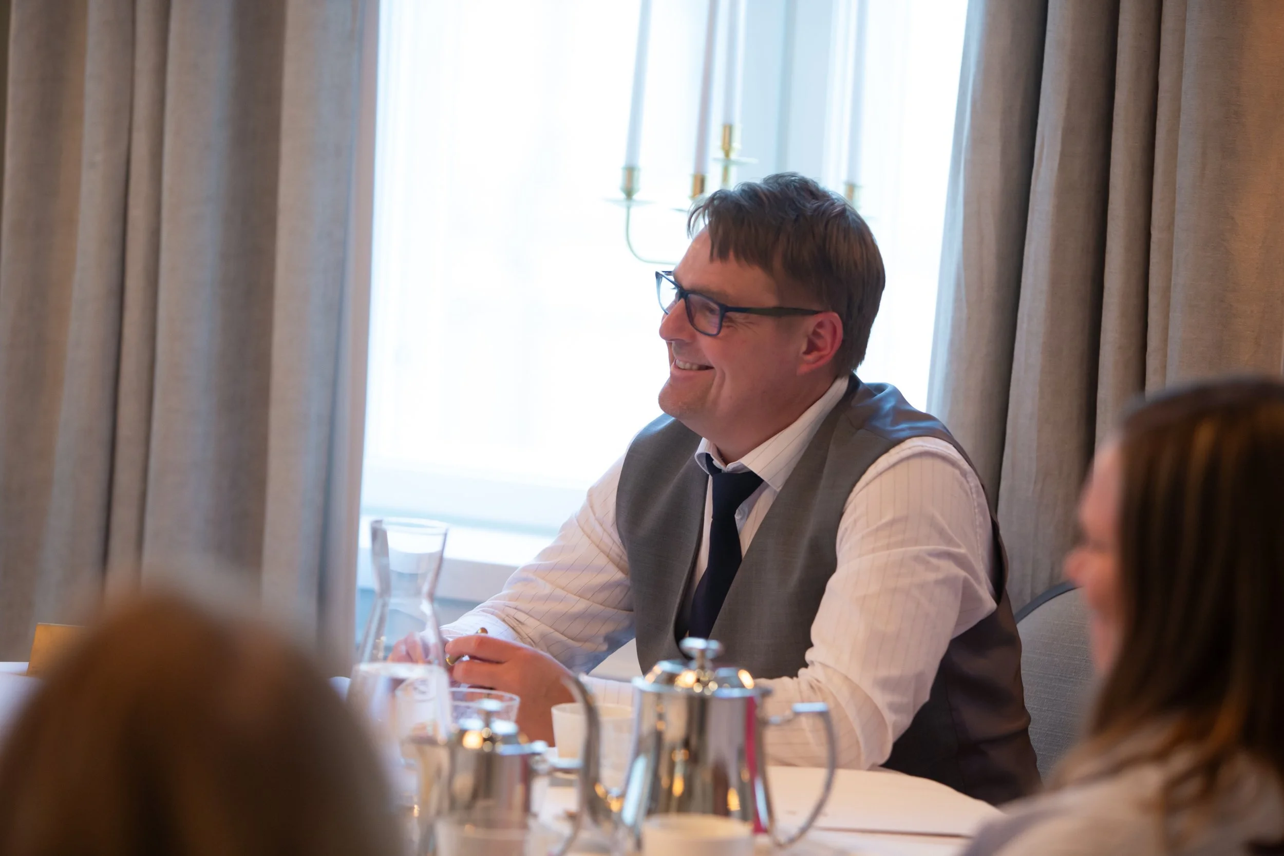 Hymyilevä mies istuu pöydässä, juhlallisessa ympäristössä, pöydällä on tarjoiluvälineitä ja tarvikkeita.