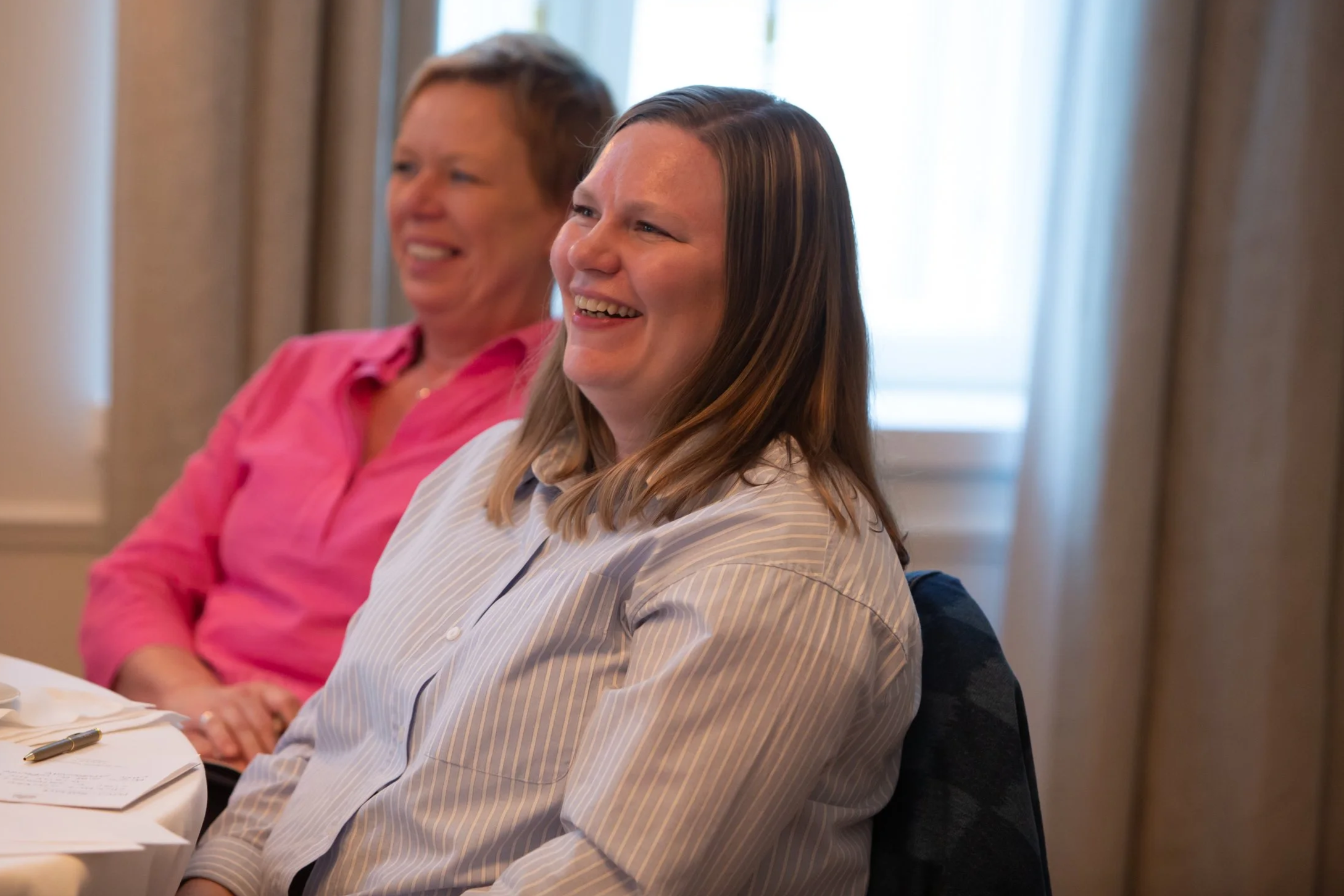 Kaksi naista istuu juhlapöydässä, nauttien huippuhetkistä, nauraen ja hymyillen.