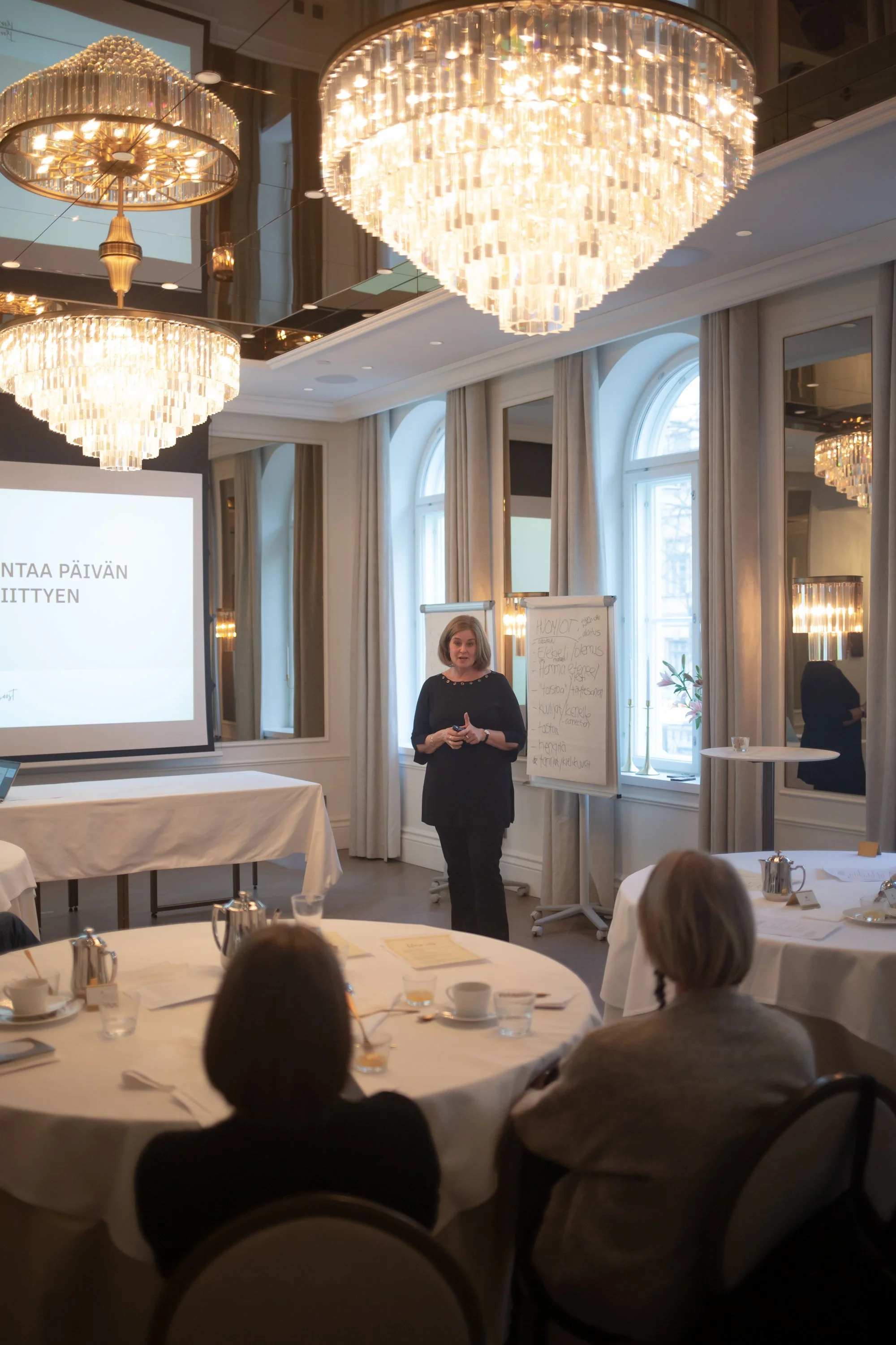 Nainen esittelee PowerPoint-esitystä ja kirjoittaa muistiinpanotaulukkoon konferenssissa tai kokouksessa, sijaitsee kauniissä ja hyvin valaistussa huoneessa juhlallisista kristallikruunuista.