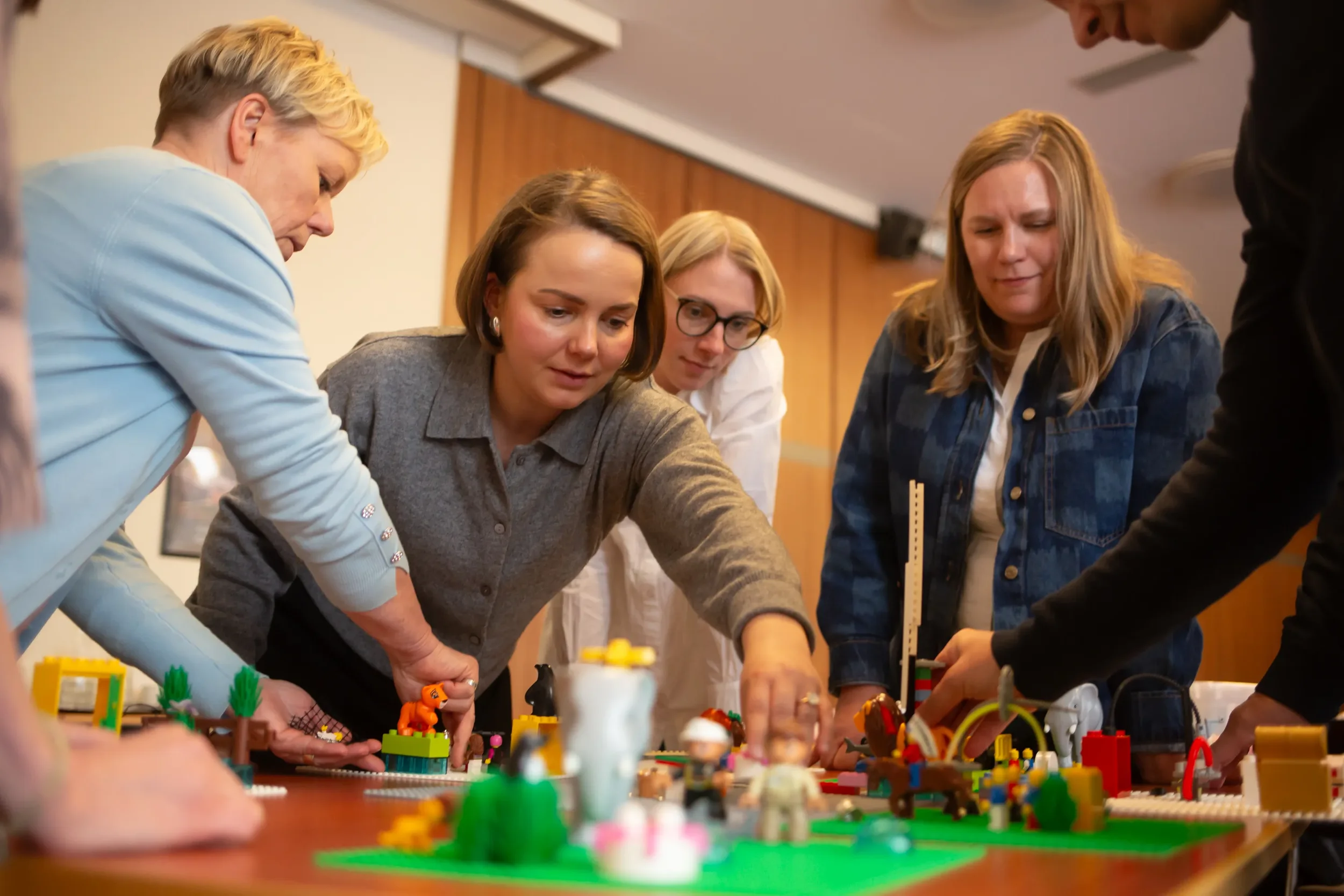 Nainen ja kolme muuta ihmistä kokoontuneet pöydän ääreen rakentamaan legoja, heillä on kiinnostus ja keskittyminen legojen rakentamiseen.