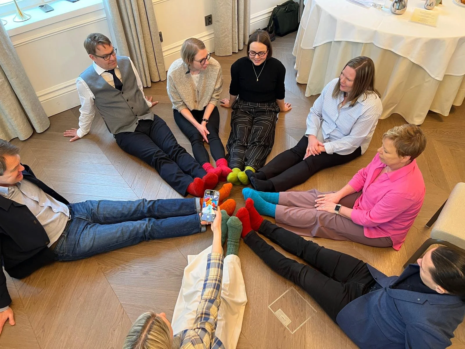 Seitsemän ihmistä istuu lattialla muodostaa ympyrän ja jokainen heistä käyttää erivärisiä villasukkia, jotka ovat keskellä heidän jalkojensa ympärillä.