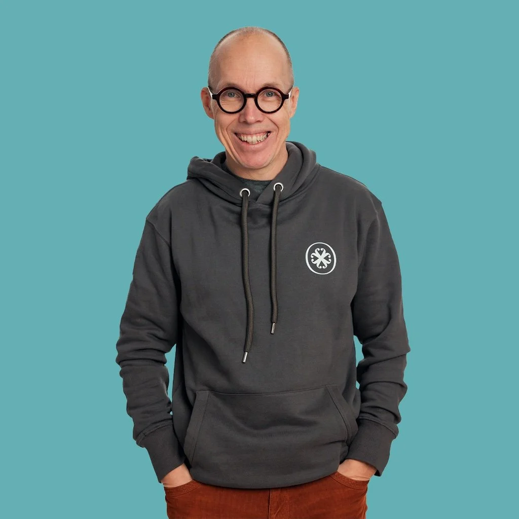 Henkilö hymyilee ja katsoo suoraan kameraan, pukeutuneena harmaaseen huppariin ja ruskeisiin housuihin, taustalla vaaleansininen väri.