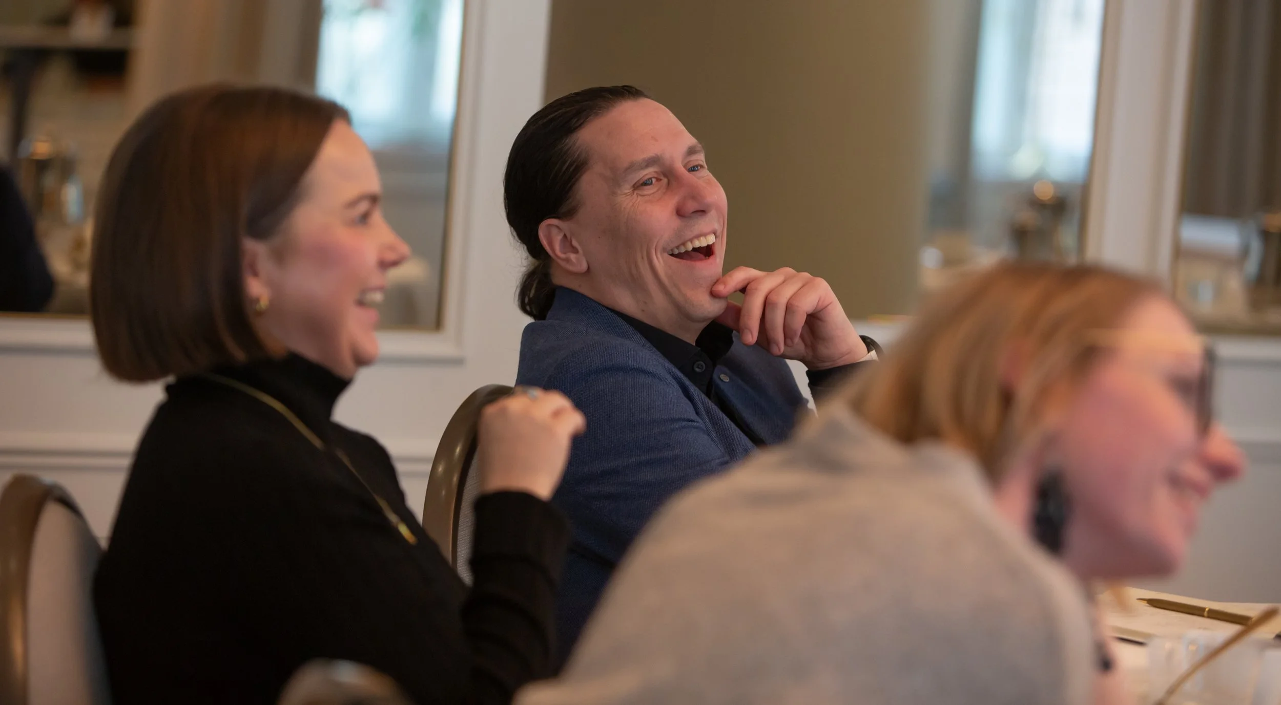 Kolme ihmistä istuu pöydän ääressä, nauravat ja katsovat jotain off-camera. Taustalla näkyy suuret peilit ja ikkunat.