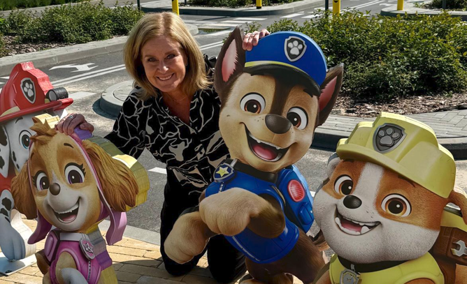 Nainen poseeraa iloisesti viiden Paw Patrol -hahmon kanssa ulkona, kaupunkialueella ja aitojen ja kivetyksen taustalla.