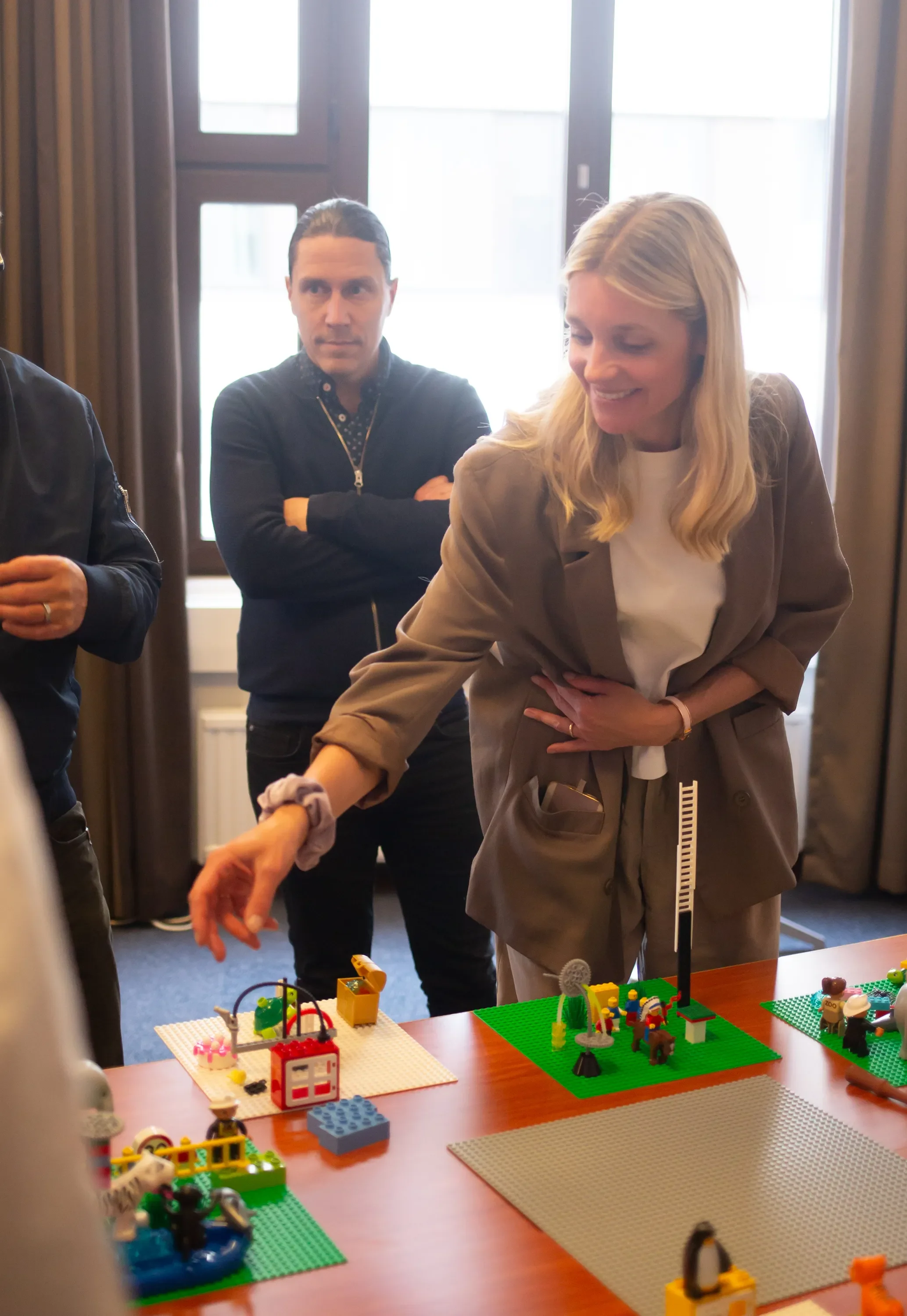 Nainen rakentaa LEGO-rakennelmaa pöydällä, kolme ihmistä katsoo sen vierestä, ikkunasta näkyy ulos.