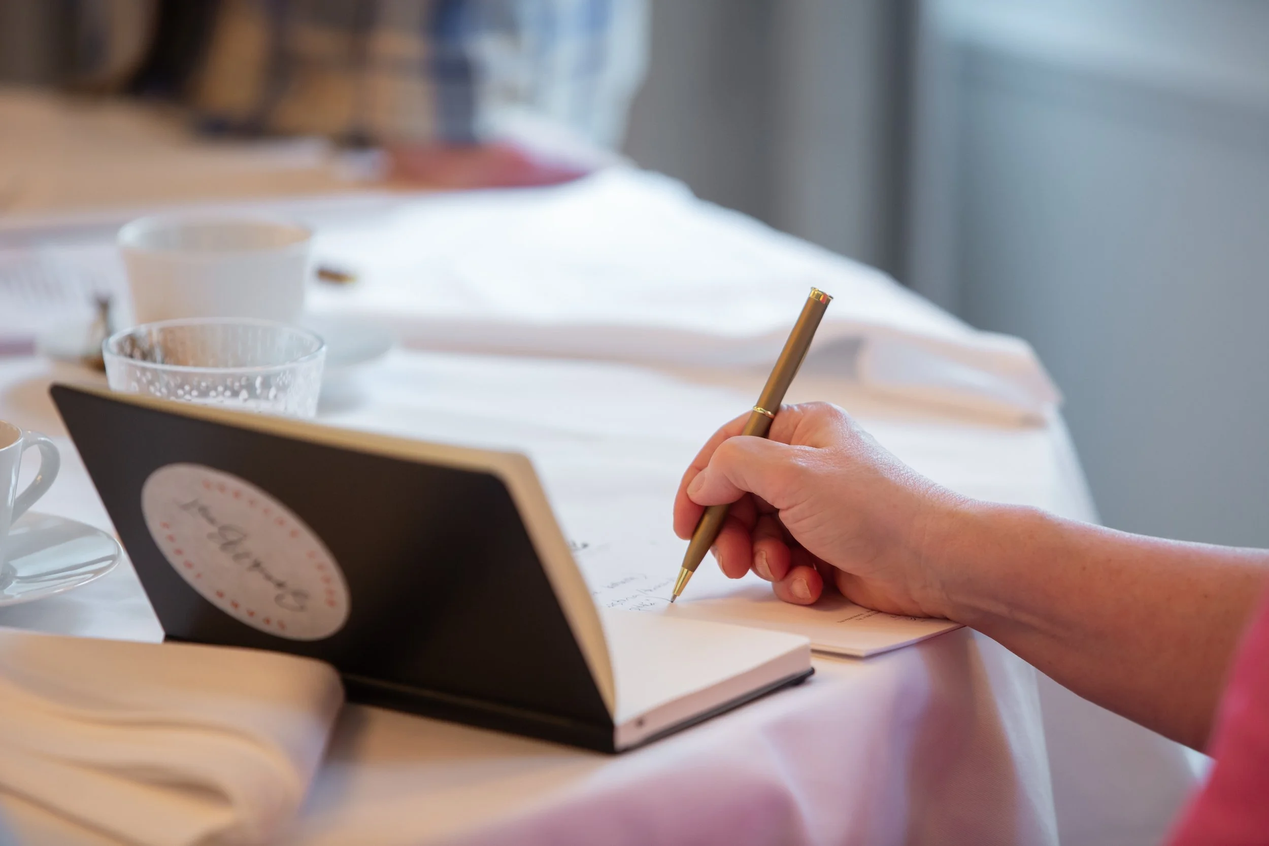 Ihmisen käsi kirjoittaa muistikirjaan, pöydällä on kahvikuppeja ja lasi.