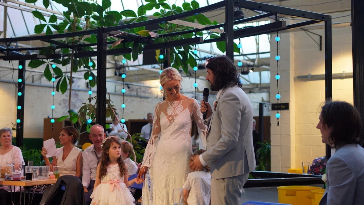 A wedding reception indoors with a bride and groom dancing and speaking into a microphone. Guests, including children and adults, watch and smile. The setting features large plants, hanging lights, and a bright, airy atmosphere.