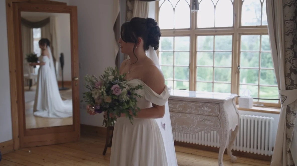 A bride in a white wedding dress holding a bouquet of flowers in front of a mirror in a bright room with large windows and wooden floors.
