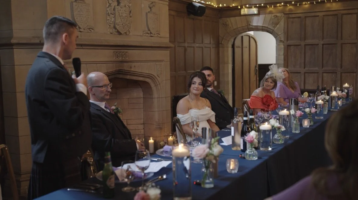 Wedding reception with a groom, bride, and guests seated at a long table with candles and flowers, a man giving a speech with a microphone.