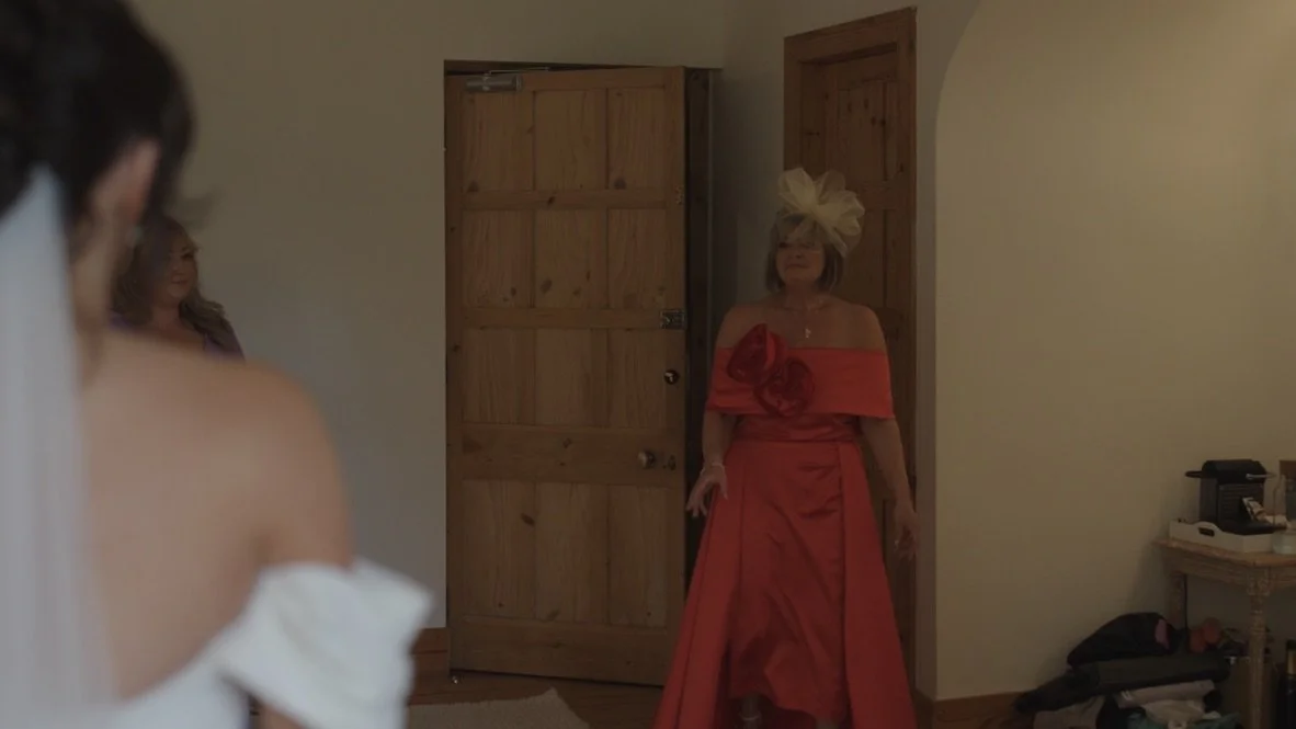 A woman in a red dress and large hat standing in a doorway, with a blurry person in a white dress in the foreground.