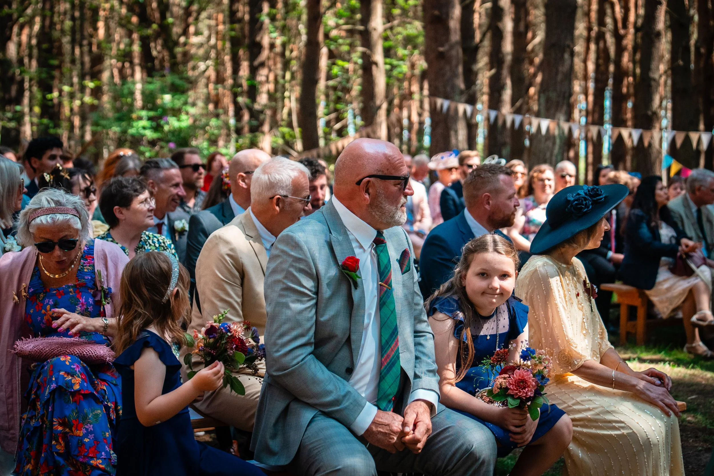 People sitting on benches outdoors in a forest, attending a wedding ceremony. The crowd includes adults and children dressed in colorful and formal attire, with some holding flowers. The background shows tall trees and bunting decorations.