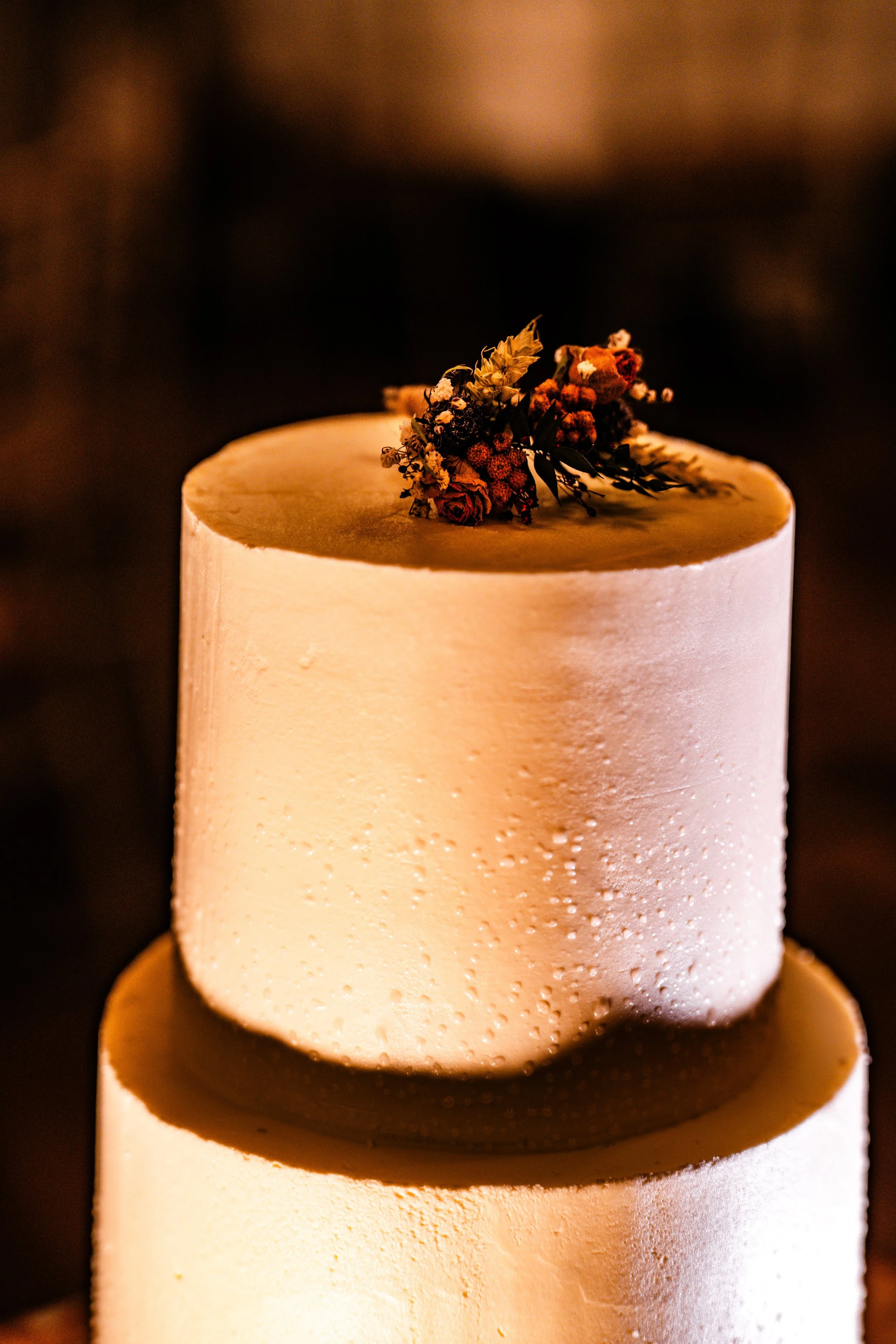 Close-up of a two-tiered wedding cake with white frosting and a small dried flower arrangement on the top tier, dark background.