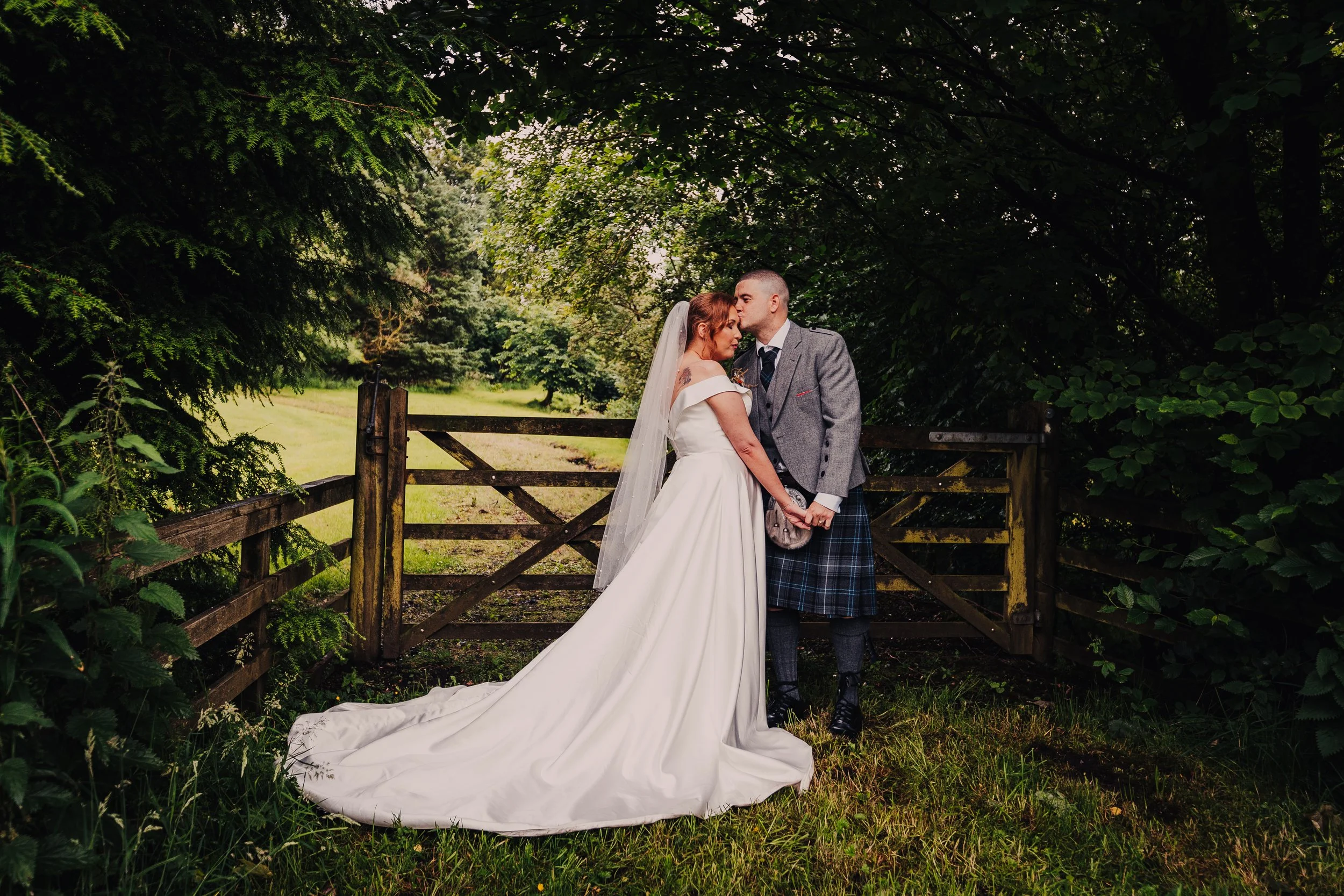 A bride and groom in wedding attire embrace in front of a wooden gate surrounded by lush green trees and grass, with the bride wearing a long white dress and veil, and the groom in a gray jacket and kilt, sharing a kiss.