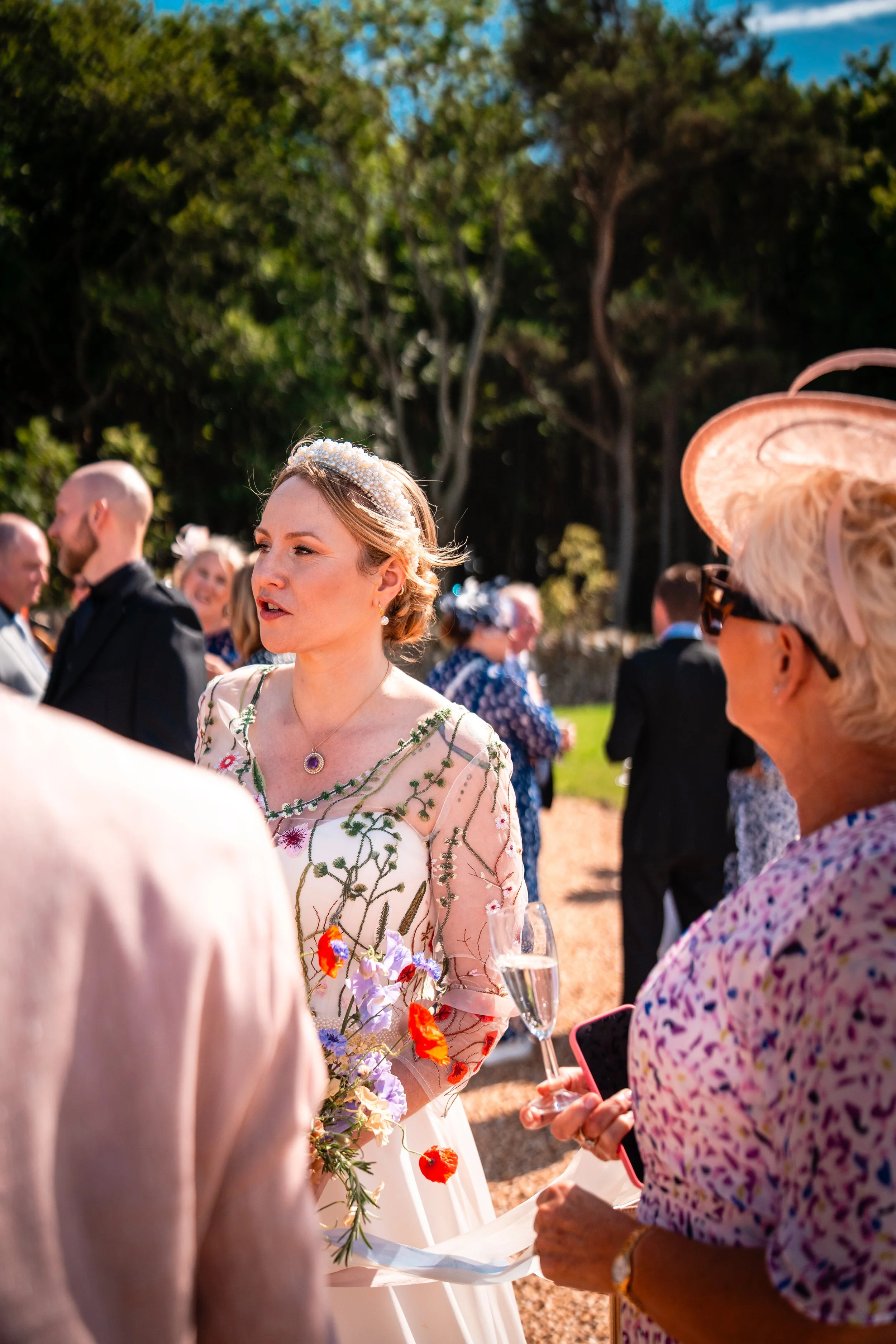 Wedding reception outdoors with guests socializing, woman in floral wedding dress holding a bouquet, woman in pink hat and sunglasses holding a glass of champagne.