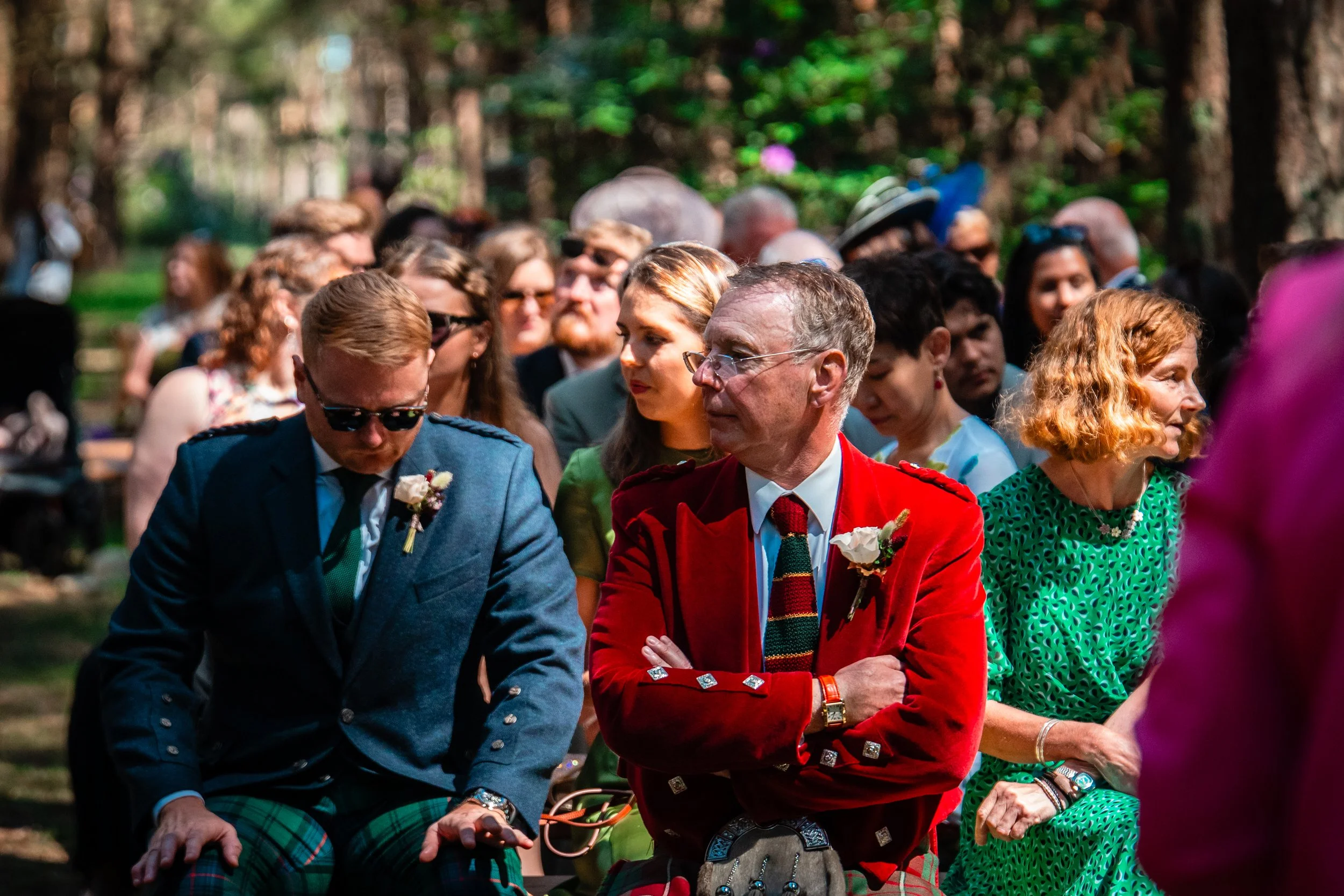 Group of people attending an outdoor event, with some wearing traditional Scottish attire, including plaid kilts, in a wooded area with sunlight filtering through trees.