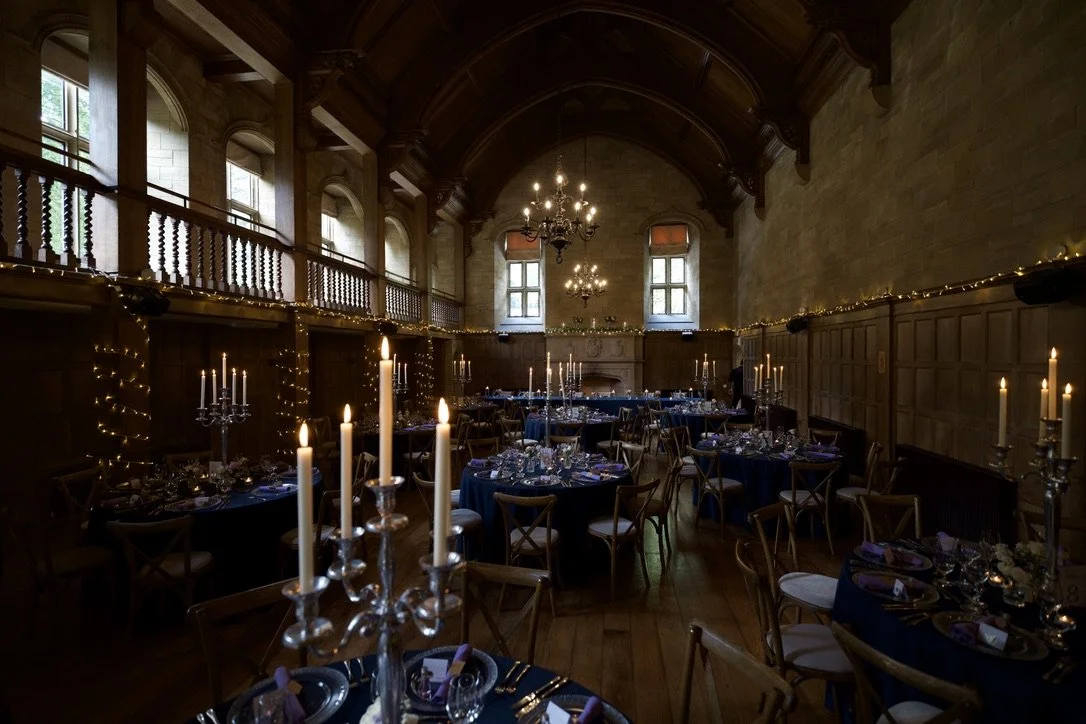 Elegant banquet hall with candles and string lights on walls, chandeliers hanging from wooden vaulted ceiling, large windows with natural light, tables set for a formal event.