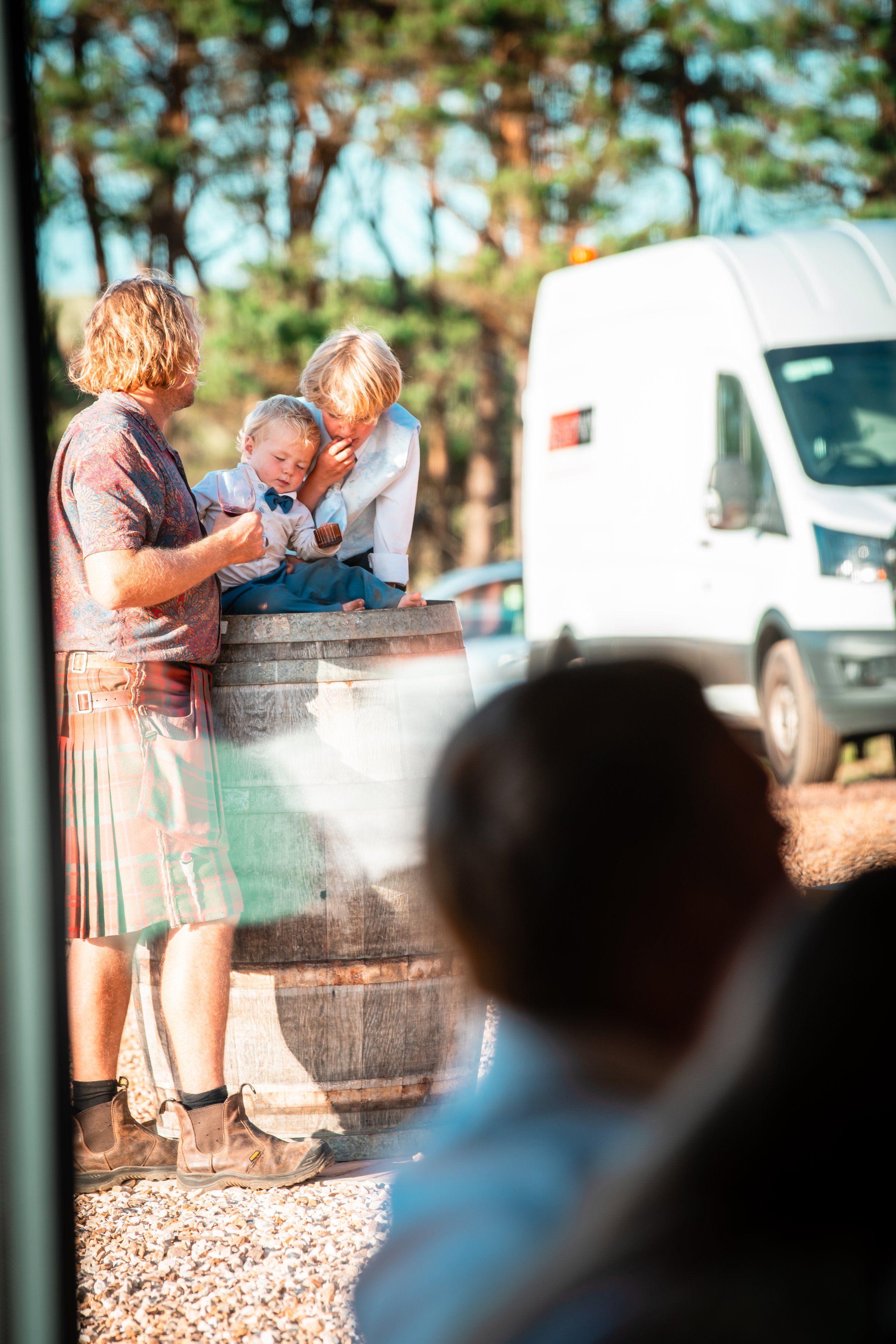 A group of three people, including a child, gathered around a barrel outdoors with trees in the background, during daytime.