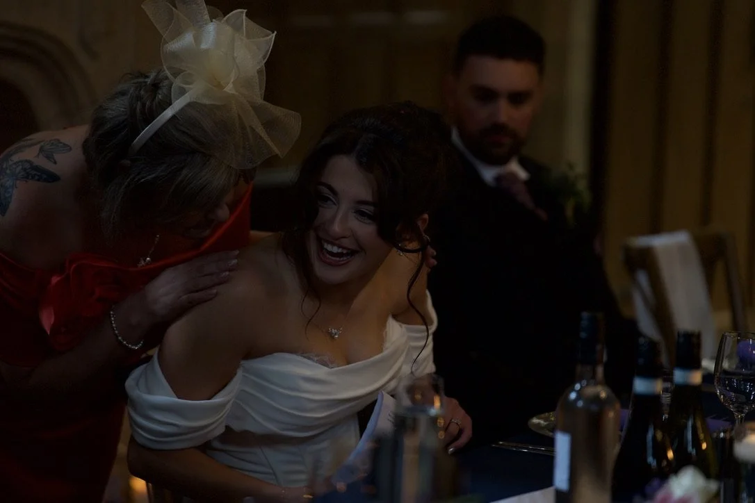 A woman in a white dress is smiling and sitting at a dinner table, talking with an older woman leaning in close with a large, decorative hair accessory. A man in a dark suit is in the background, observing the interaction. The setting appears to be a