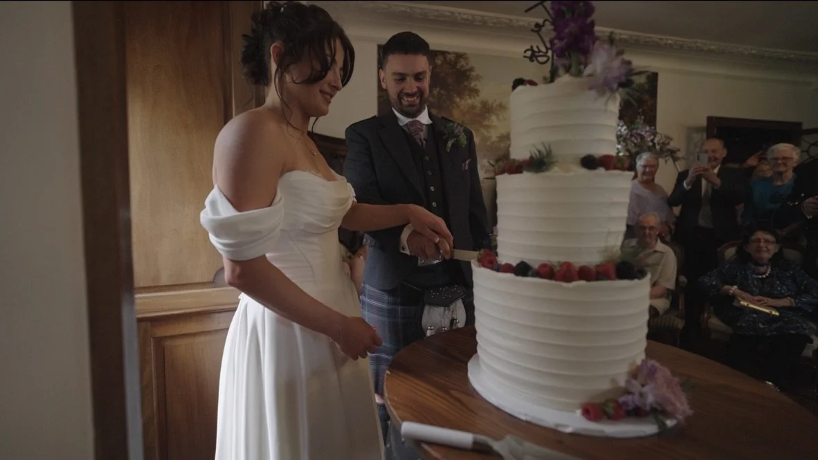A bride and groom cutting a multi-tiered wedding cake decorated with berries and flowers, with guests watching and smiling in the background.