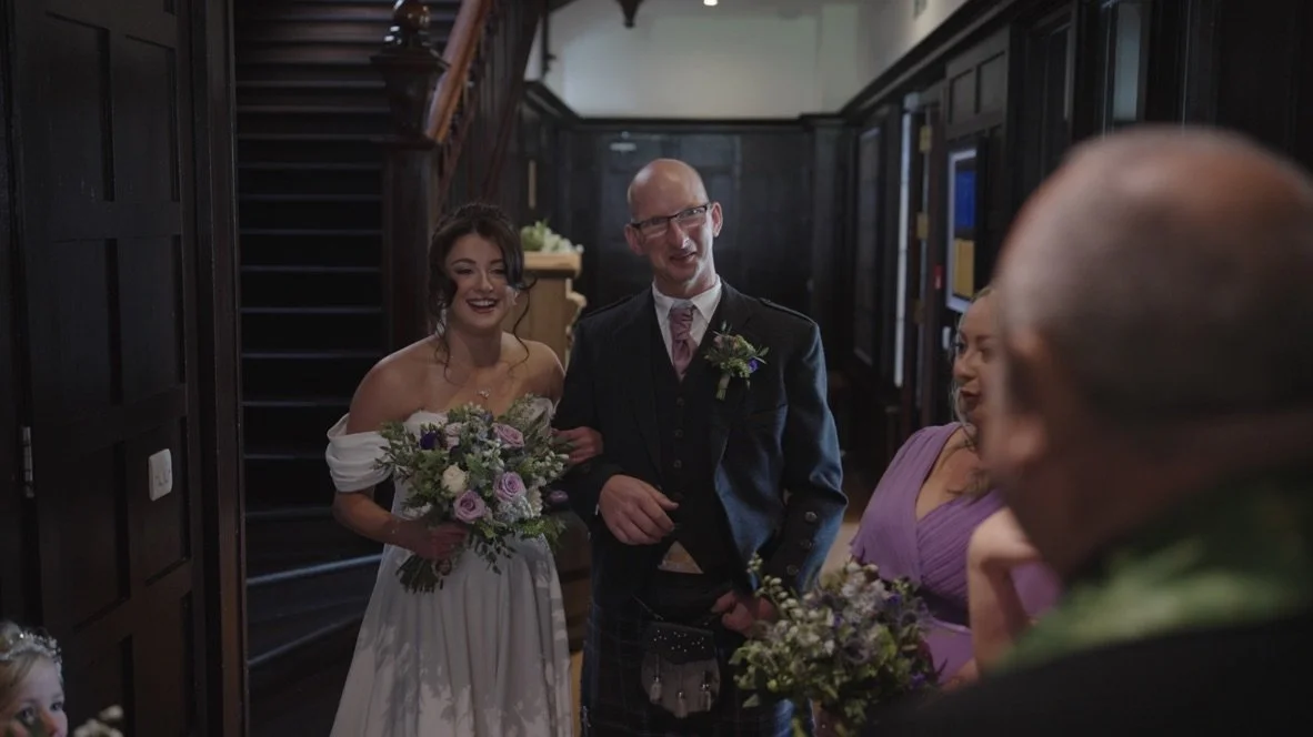 People at a wedding reception, including a bride in a white dress holding a bouquet, a man in a suit and kilt, and women in purple dresses, smiling and enjoying the moment in a wood-paneled room.