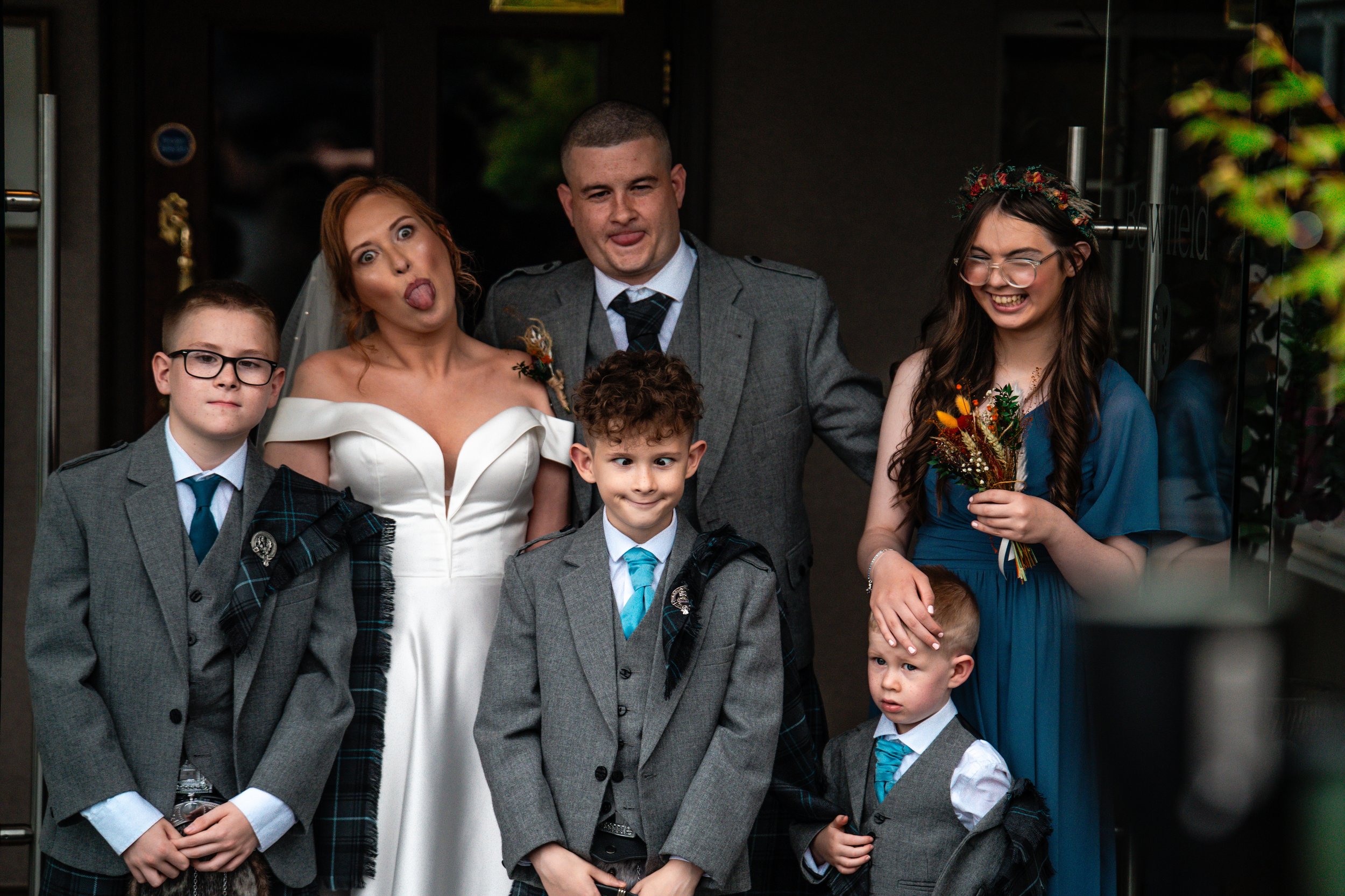 Group of seven people, including children and adults, at a wedding, making playful facial expressions. The woman in an off-shoulder white dress, the man in a gray suit, and women in blue dresses are smiling or sticking out their tongues, standing out