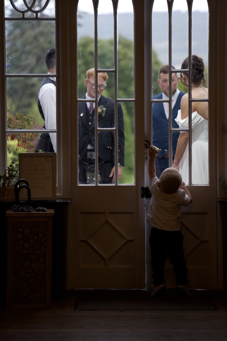 A young boy standing indoors holding the door open for a wedding procession outside, where a bride and three groomsmen are visible through the glass panes of the door.