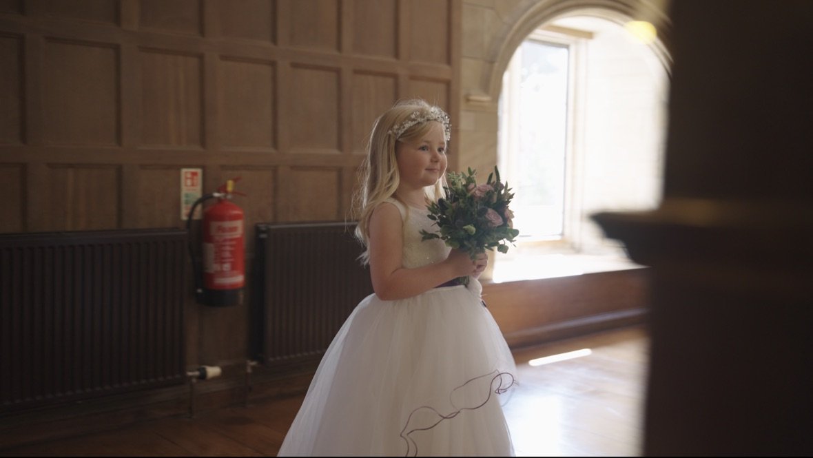 A young girl in a white dress holding a bouquet of flowers, standing indoors in a room with wooden walls and large arched window.