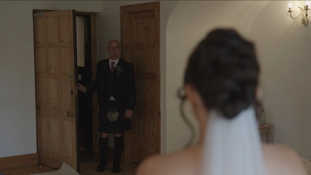 A groom standing in a doorway looking at a bride who is blurred in the foreground, with wooden doors and a wall-mounted light fixture in the background.