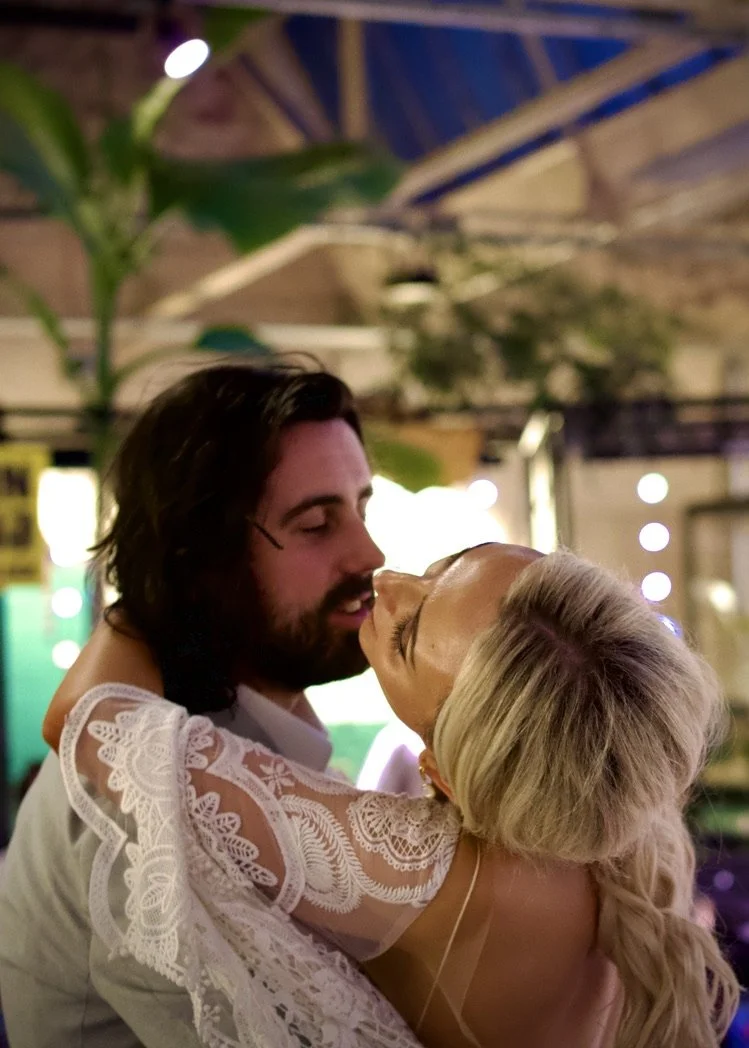 A couple sharing a kiss indoors, with the woman wearing a white lace dress and having blonde hair, and the man with dark hair and a beard.