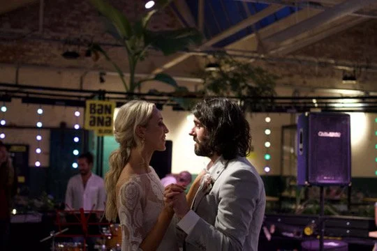 A man and woman holding hands and looking at each other at a party or event, with colorful lights and other people in the background.