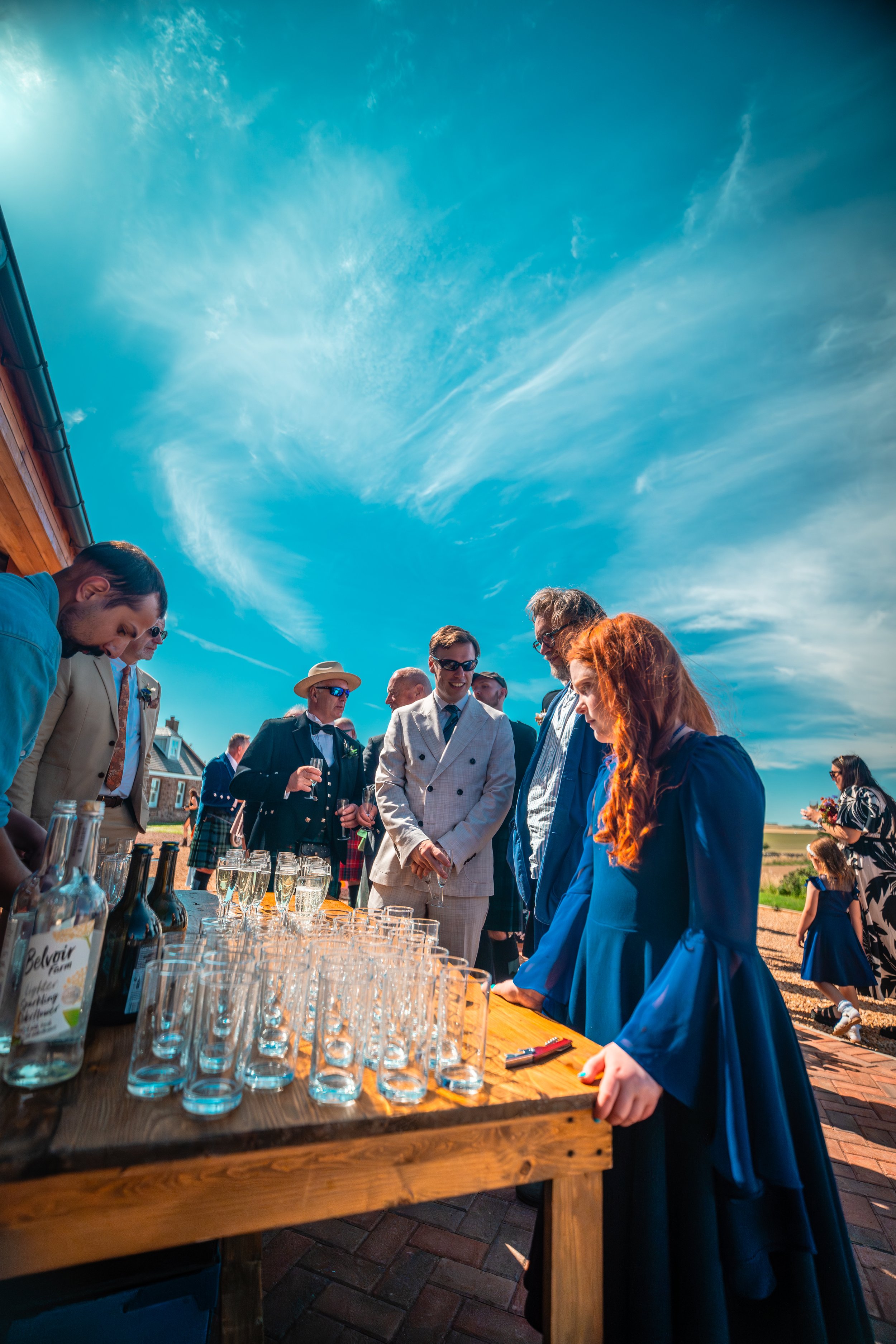 People at an outdoor celebration around a wooden table with glasses and bottles against a bright blue sky.