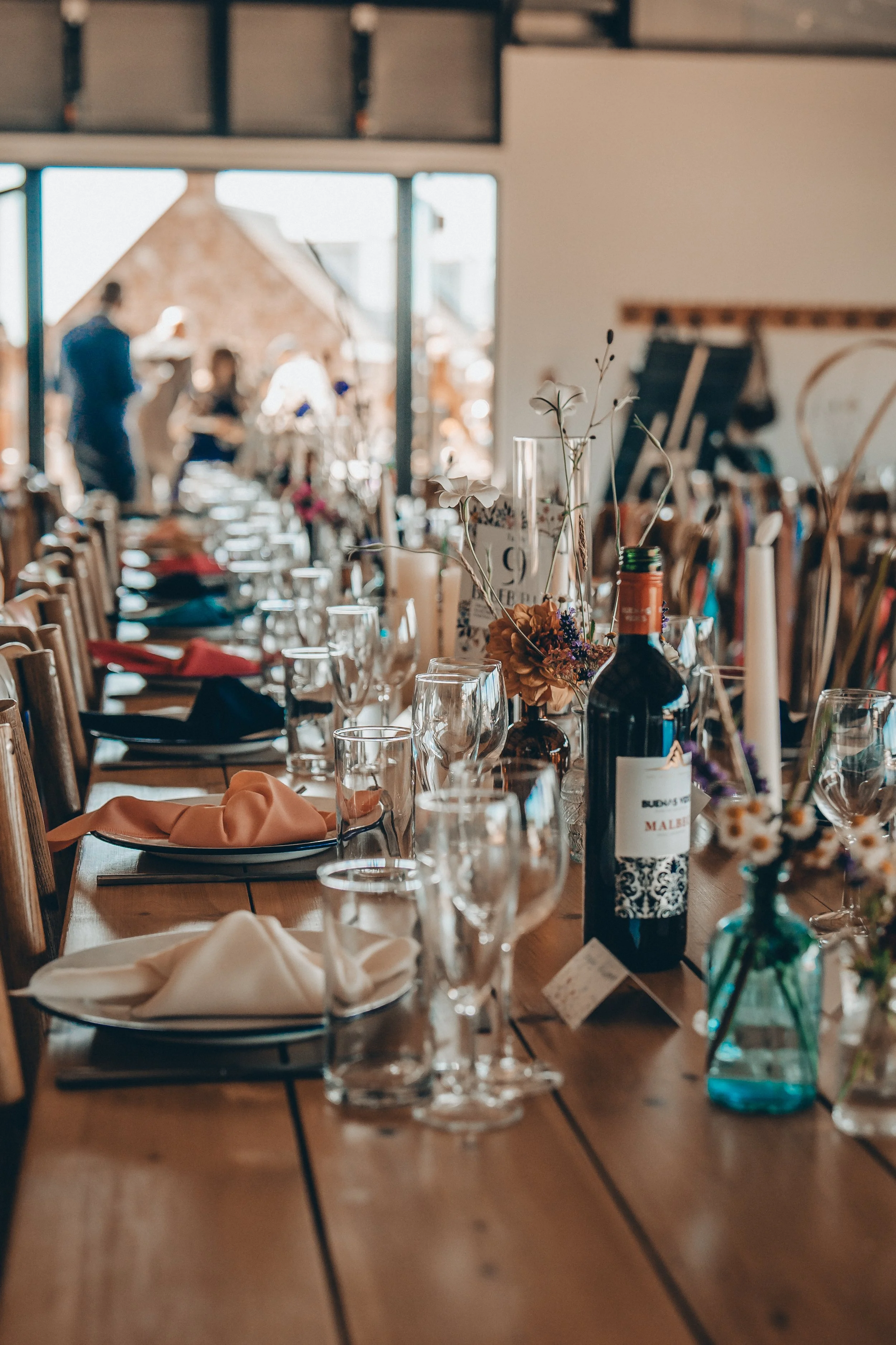 Elegant table setup with wine bottles, candles, flowers, and glassware in a warmly lit dining area with people talking in the background.