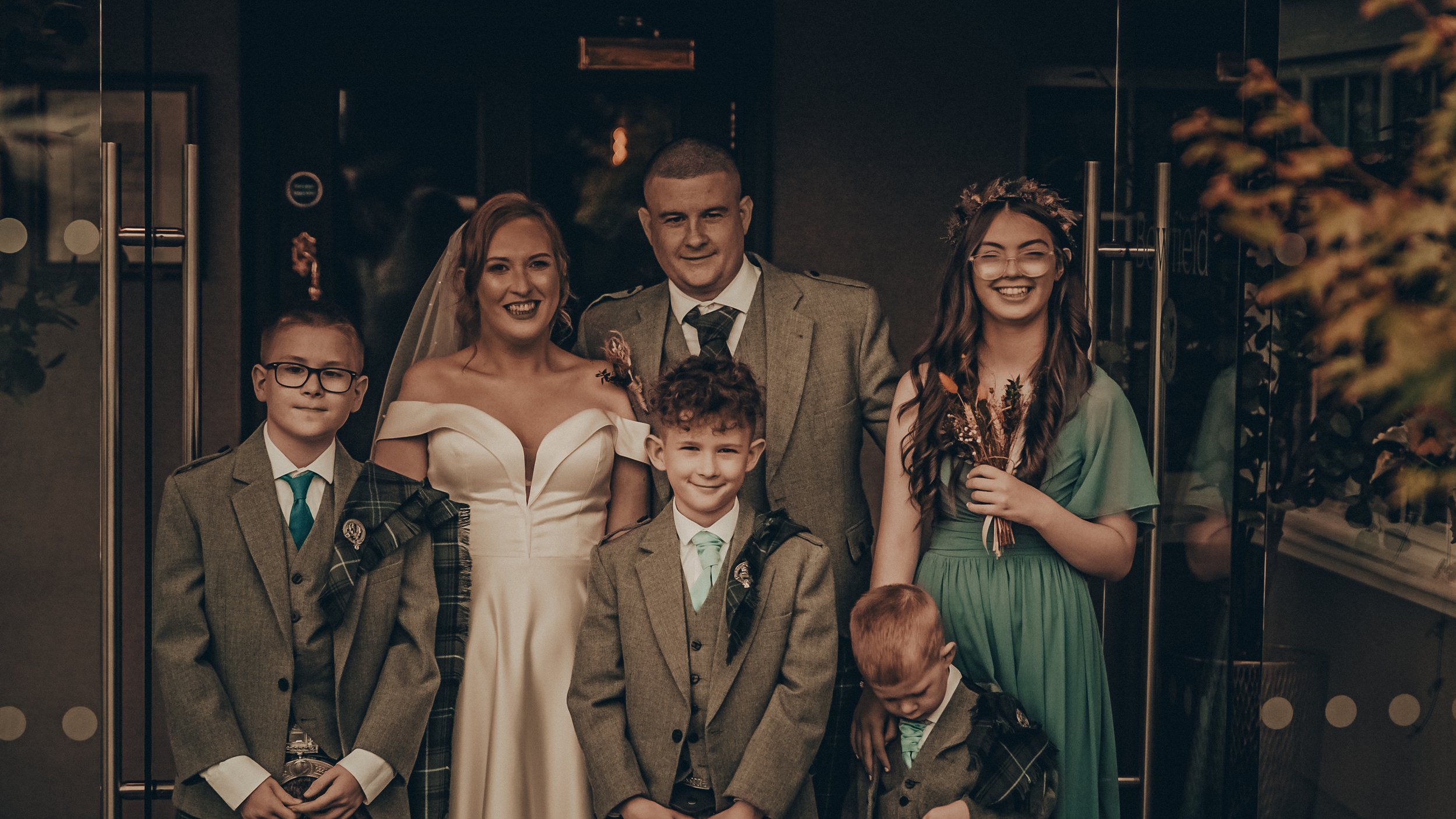 Family at wedding, including a bride, groom, three young boys in suits, and a young woman in a green dress holding a bouquet, standing together indoors in front of glass doors.