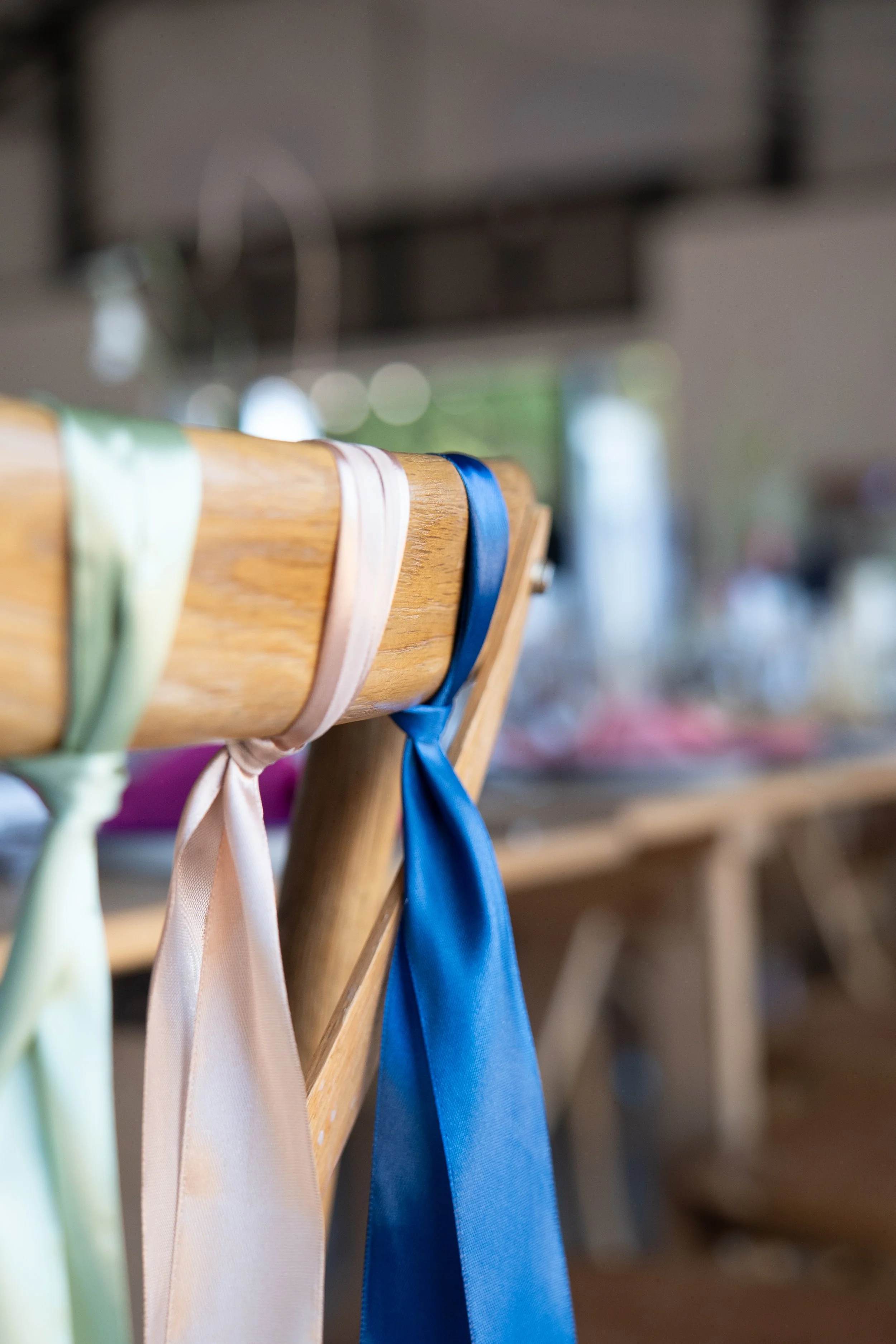Close-up of a wooden chair with colorful satin ribbons tied to each chair's backrest, including pink, cream, and blue ribbons, in a setting that appears to be prepared for an event.