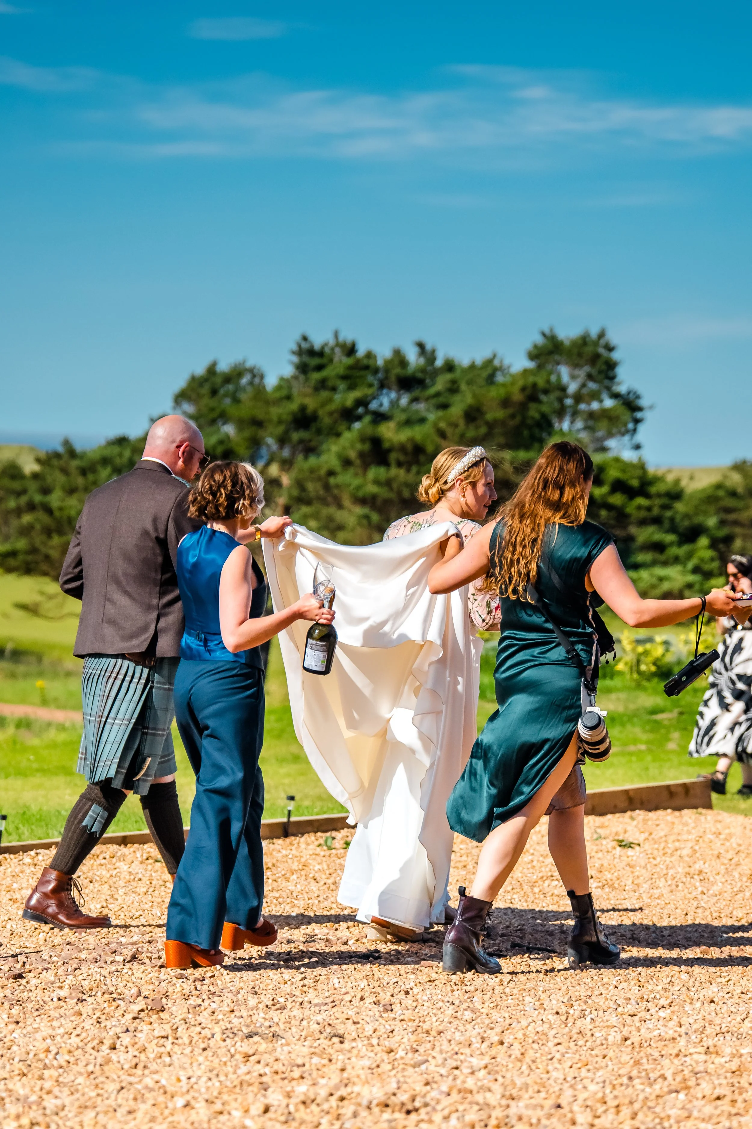 People at a wedding ceremony outdoors on a sunny day, with a bride in a white dress, accompanied by others, on a grassy area with trees in the background.