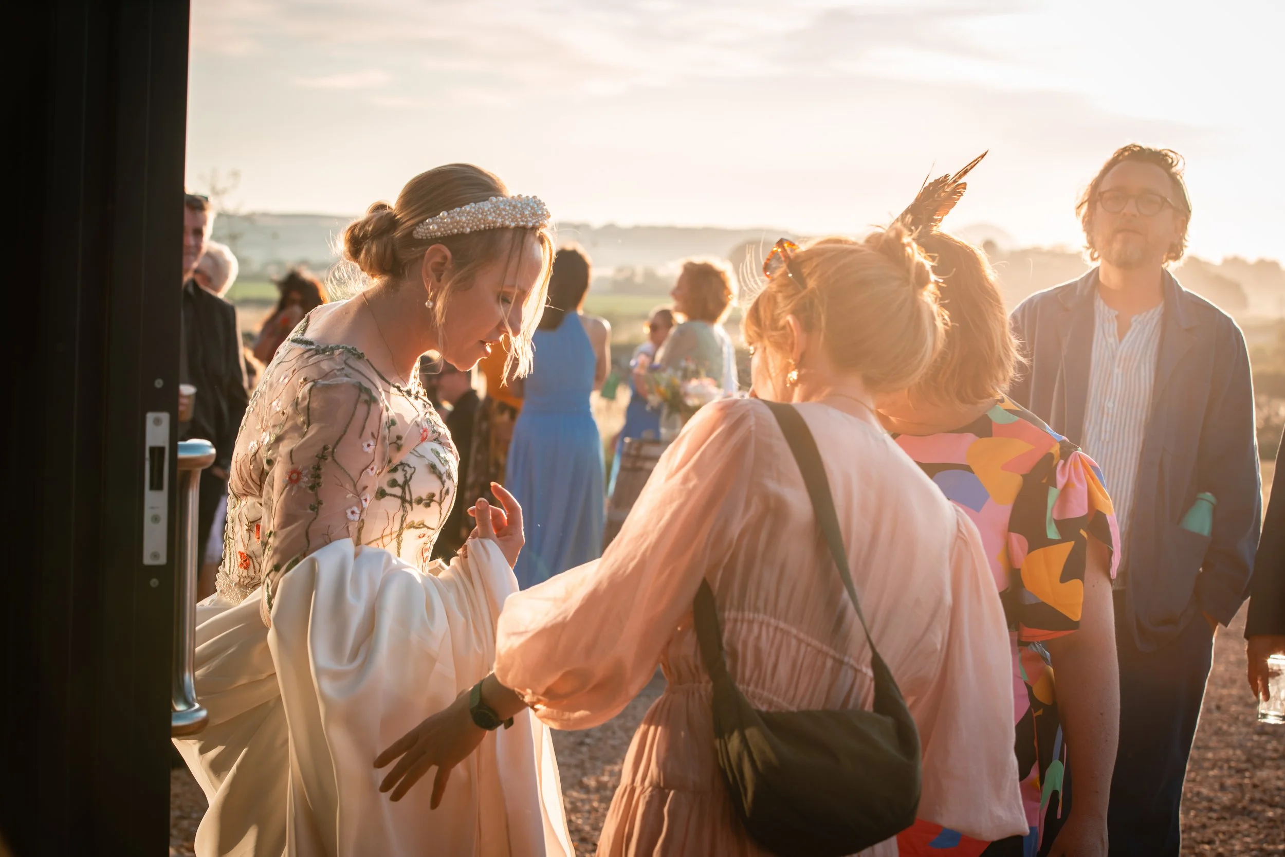 A woman in a floral dress and pearl headband is smiling and talking to a group of people outdoors during sunset, with rolling hills in the background.