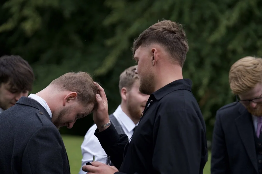 Group of men in formal attire outdoors, engaged in a serious or emotional moment, with one man touching another's head.