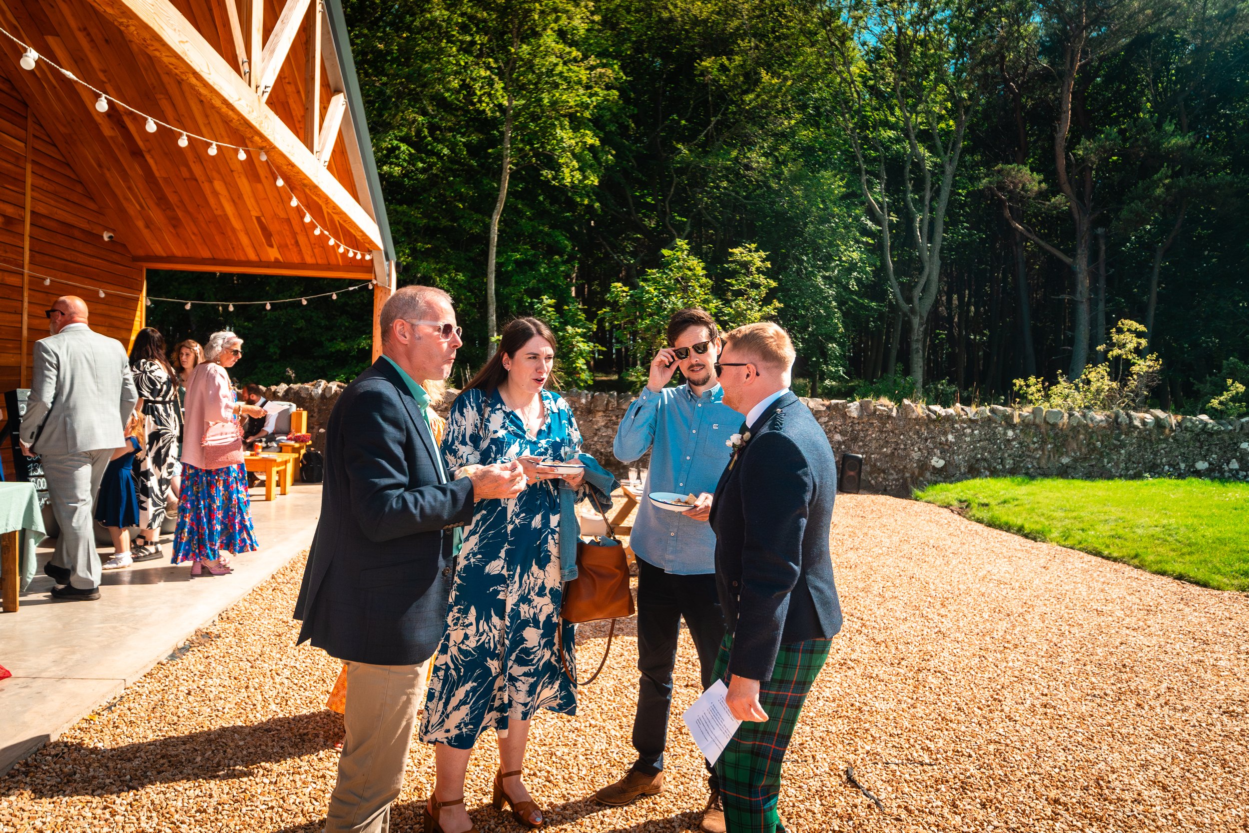 Group of people at an outdoor event, socializing on a sunny day with a wooden building and trees in the background.