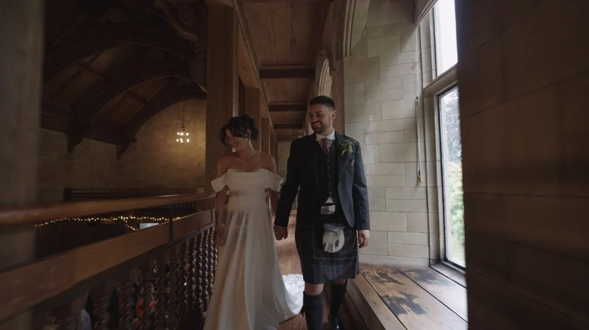 A bride in a white wedding dress and a groom in a dark suit holding hands and walking down a wooden corridor inside a church or hall with natural light coming through a window.