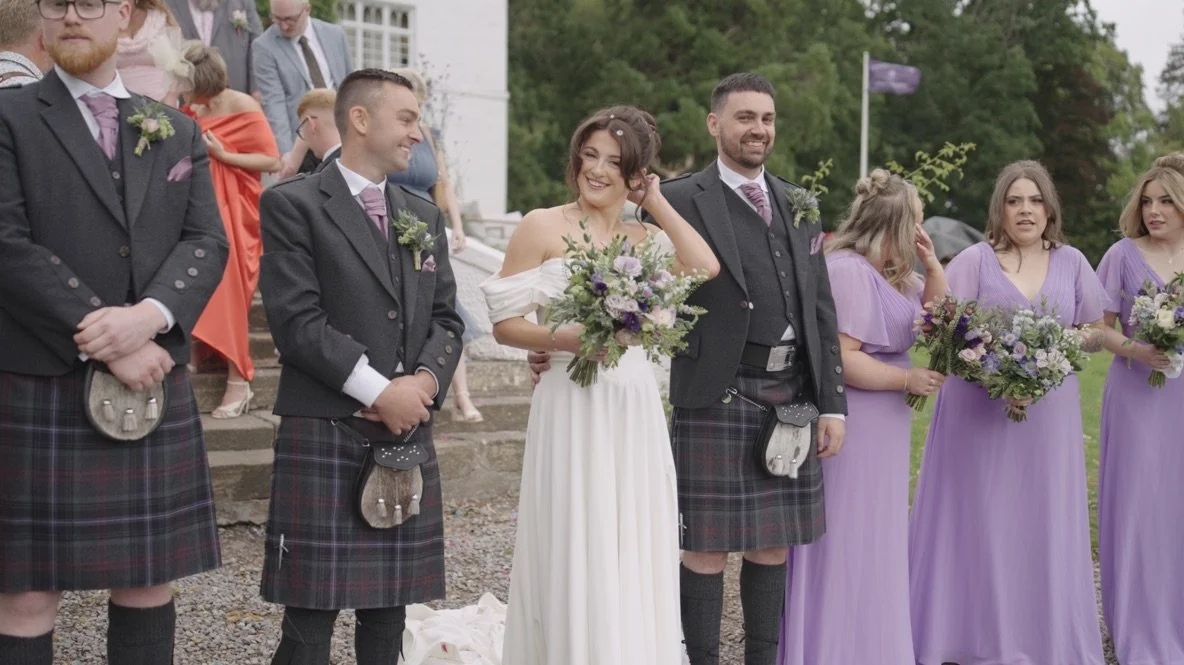 A bride and groom with men and women dressed in formal attire, outdoors at a wedding. The bride is holding a bouquet, and the women are wearing lavender dresses, while the men wear traditional Scottish kilts and jackets.