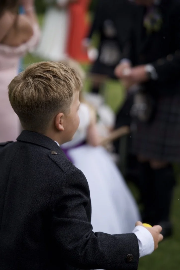A young boy with blond hair wearing a black blazer, holding a yellow object, at an outdoor event with other children and adults in the background.