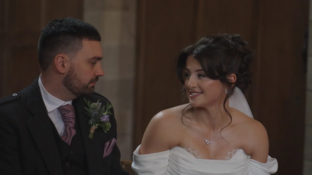 A bride and groom sit together during a wedding ceremony, smiling at each other in a warmly lit church setting.
